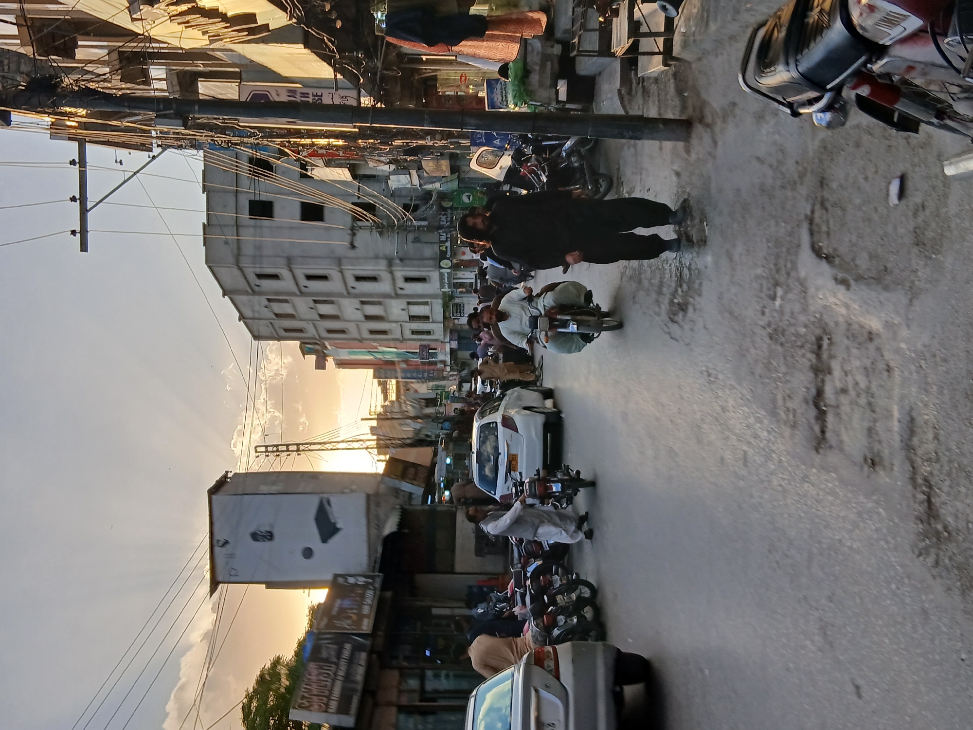 The image depicts a vibrant street scene in an urban area, likely Quetta City Tehsil, Pakistan, during either sunrise or sunset. The sky, bathed in a warm, golden light, shows visible sun rays emanating from behind buildings or clouds towards the left side of the frame.
The street is bustling with daily activity. Numerous people are present; a prominent man in a black shalwar kameez walks on the pavement towards the right foreground. Other pedestrians are visible further down the street. Many men are riding or standing by motorcycles, including one in a white shalwar kameez and waistcoat on a motorcycle approaching the foreground. Several cars are also visible, a white sedan and a silver car being most distinct.
Multi-story buildings, some appearing residential or commercial, line both sides of the road. One building in the mid-ground features a grid-like pattern on its facade. Overhead, a dense and complex network of utility poles and tangled electrical wires crisscrosses the scene, a common feature in urban South Asia. Various commercial signs and billboards are visible, though most text is unreadable due to distance or angle. A building sign in the mid-ground partially displays "AL HA...". The asphalt road shows signs of wear and tear, with some debris visible. The overall atmosphere is that of a busy urban environment captured during the golden hour.