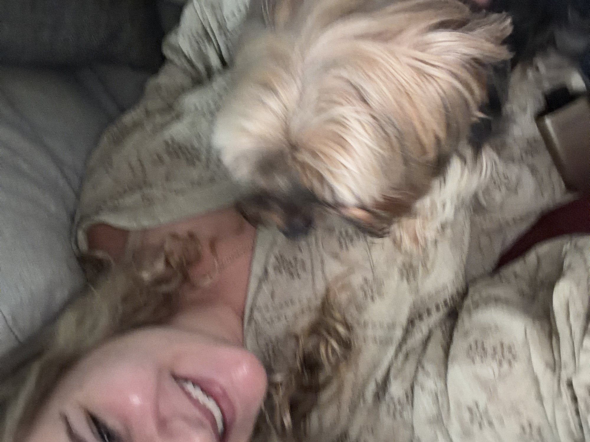 A woman with blonde hair is smiling in the lower left of the frame. A small, light brown dog is resting on her chest. She is lying on a light colored patterned blanket. The location, San Bernardino Tlaxcalancingo, Mexico, is not evident from the image.