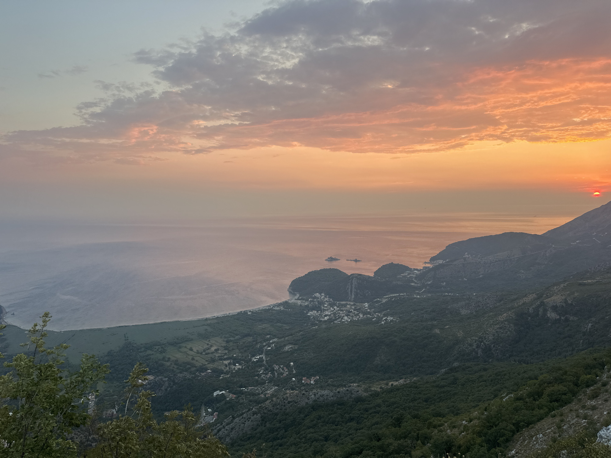 The image captures a panoramic view from a high vantage point overlooking the Adriatic Sea and the coastal region near Gluhi Do, Montenegro. The scene is bathed in the warm, diffused light of a sunset, with the sky displaying a gradient of soft blues, pinks, and oranges, tinged with scattered clouds. The sun, a small, bright red disc, is partially visible on the horizon to the right.
Below the sky, the calm sea stretches out, its surface reflecting the sky's colors in muted tones. Along the coastline, a crescent-shaped beach is visible, bordered by a stretch of green land that transitions into lush, dense forest covering the rolling hills and mountains. The terrain is rugged, with steep inclines and shadowed valleys.
Nestled within the landscape, small clusters of buildings, indicative of a village or town, are scattered across the slopes and near the coast. Roads wind through the greenery, connecting these settlements. In the foreground, the tops of dark green trees and bushes are visible, framing the expansive vista. The overall impression is one of natural beauty and tranquility at dusk.