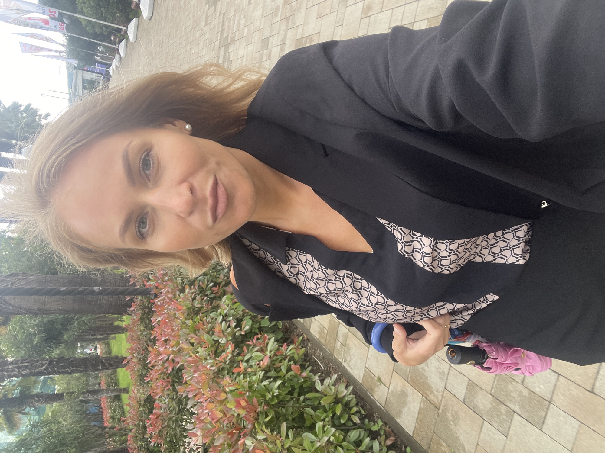 A woman is taking a selfie outdoors, dressed in a black blazer over a black and beige patterned top. She has blonde hair, fair skin, and is wearing an earring. She is holding a closed, colorful umbrella with a pink cover. The background shows a paved walkway and greenery, with trees and flags visible further back. Based on the style of the flags and architecture, the location is likely Sochi, Russia.