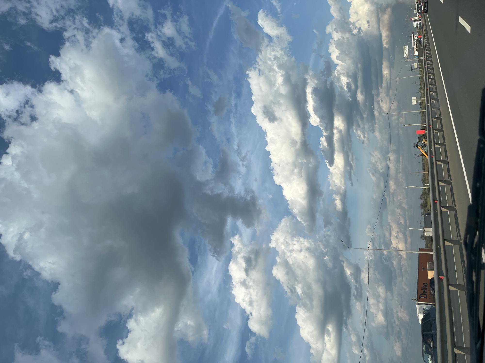 The image depicts a view from a vehicle traveling on a multi-lane highway. The sky is filled with dramatic cumulus clouds, ranging from bright white to dark gray, against a backdrop of vivid blue sky. On the left side of the highway, there is a large orange truck with the logo "Delko" visible. In the distance, there are power lines, streetlights, and some buildings or structures, suggesting an urban or industrial area. A yellow excavator is also present on the left side of the road, near some construction barriers. Vehicles are seen on the highway in both directions. The foreground shows the dashboard of the vehicle from which the photo is taken. The scene is set in Bartolomeevskii, Russia, indicated by the presence of the "Delko" truck which is a Russian logistics company.
