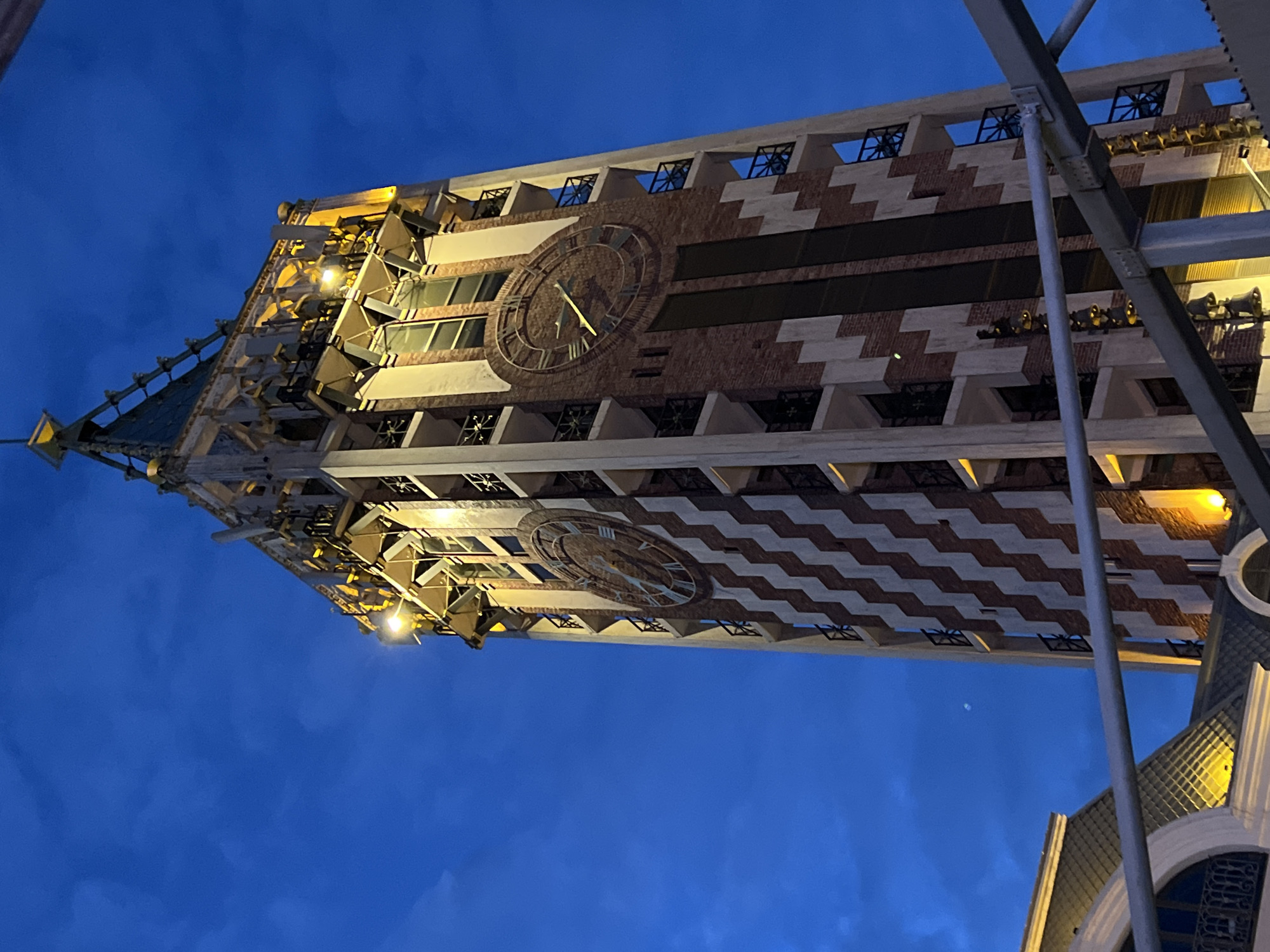 This image captures the majestic clock tower of the Batumi Piazza in Batumi, Georgia, during the evening or twilight hours. The tower stands tall against a deep indigo-blue sky, which hints at the fading daylight or the early stages of night.
The primary subject is the ornate tower, illuminated by warm yellow lights that highlight its architectural details. The building's facade features a striking pattern of reddish-brown brickwork alternating with lighter, possibly stone or stucco, sections arranged in a chevron or zigzag design. Several large clock faces are visible, one prominently in the center of the image, displaying Roman numerals and golden hands. The time on this clock appears to be around 4:50, with the minute hand pointing towards the X and the hour hand just past the IV.
Towards the top of the tower, an intricate spire is visible, along with multiple openings that might house bells. Numerous golden, horn-like structures, characteristic of a carillon, are arranged in rows along the upper sections of the tower, suggesting it plays musical chimes. Below these, there are several windows, some adorned with decorative grilles.
In the foreground, on the right side, there are metal structural elements, possibly part of scaffolding or an adjacent building, partially obscuring the tower. The upward perspective emphasizes the height and grandeur of the architecture. The sky is largely clear, with some subtle cloud formations visible. There are no people visible, and no discernible text can be read on the building. The scene conveys a sense of serene evening beauty and architectural splendor.
