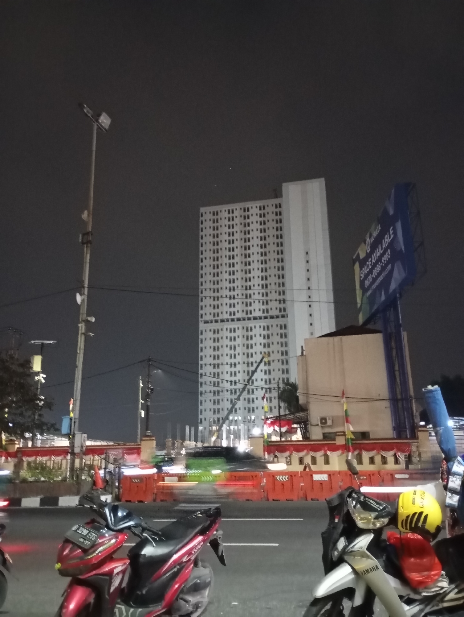 The image depicts a nighttime street scene in Depok, Indonesia, likely on a major thoroughfare. In the background, a tall, modern apartment building dominates the skyline, its facade covered in a grid of windows. To its right, a large billboard displays the text "SPACE AVAILABLE" and a phone number, indicating a commercial or residential development. The foreground features several parked motorcycles, suggesting a public area or a parking spot along the road. Traffic lights are blurred due to motion blur, indicating the presence of moving vehicles. The street is illuminated by streetlights and the lights from the buildings, creating a characteristic urban nighttime ambiance. The presence of construction barriers and flags suggests ongoing development or a local event.