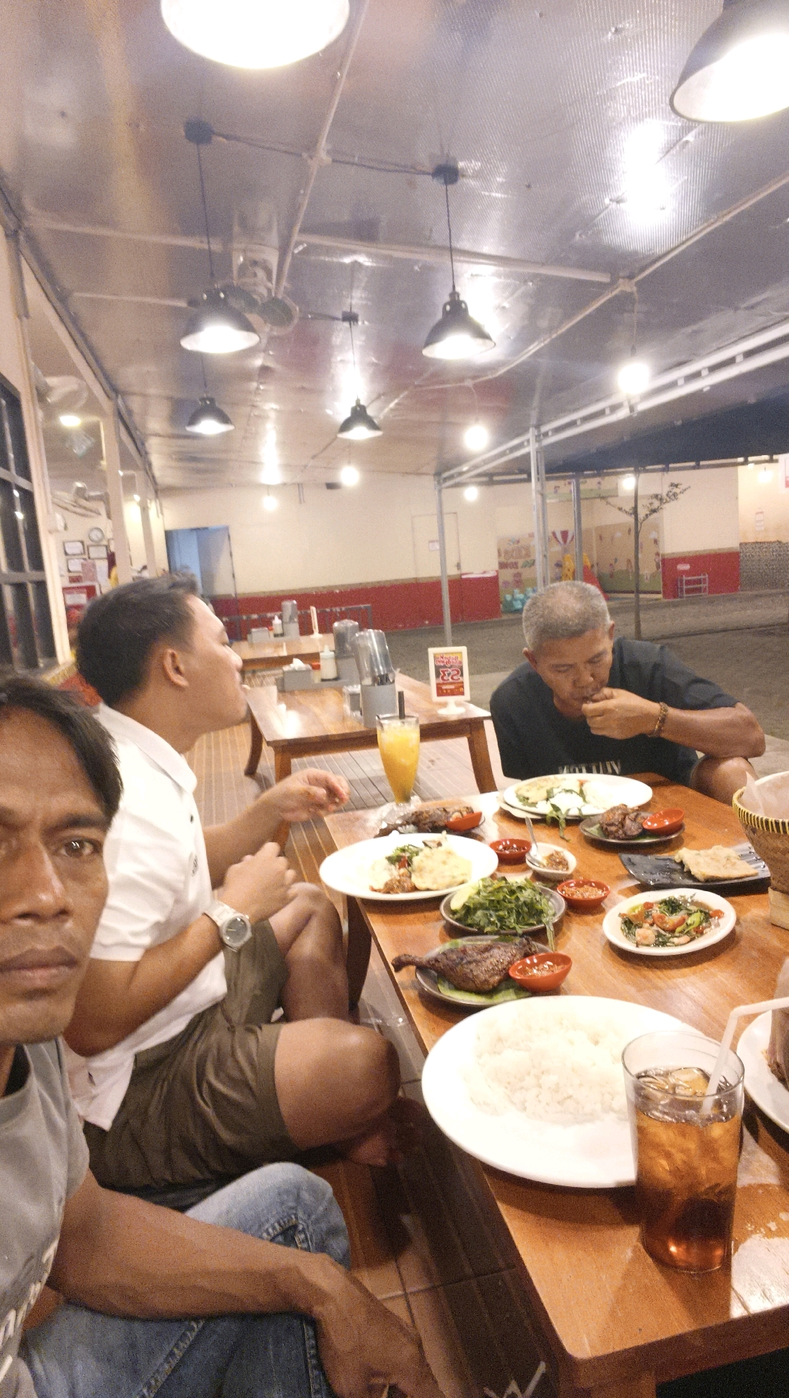 A group of men are dining at an outdoor restaurant in Ketapang, Indonesia. The setting appears to be a casual eatery with wooden tables and chairs. Several plates of food, including rice, vegetables, and grilled meat, are spread across the tables. One man is actively eating, while others are engaged in conversation or observing the food. The lighting suggests it is evening. The background shows the restaurant's structure and a glimpse of the surrounding area.