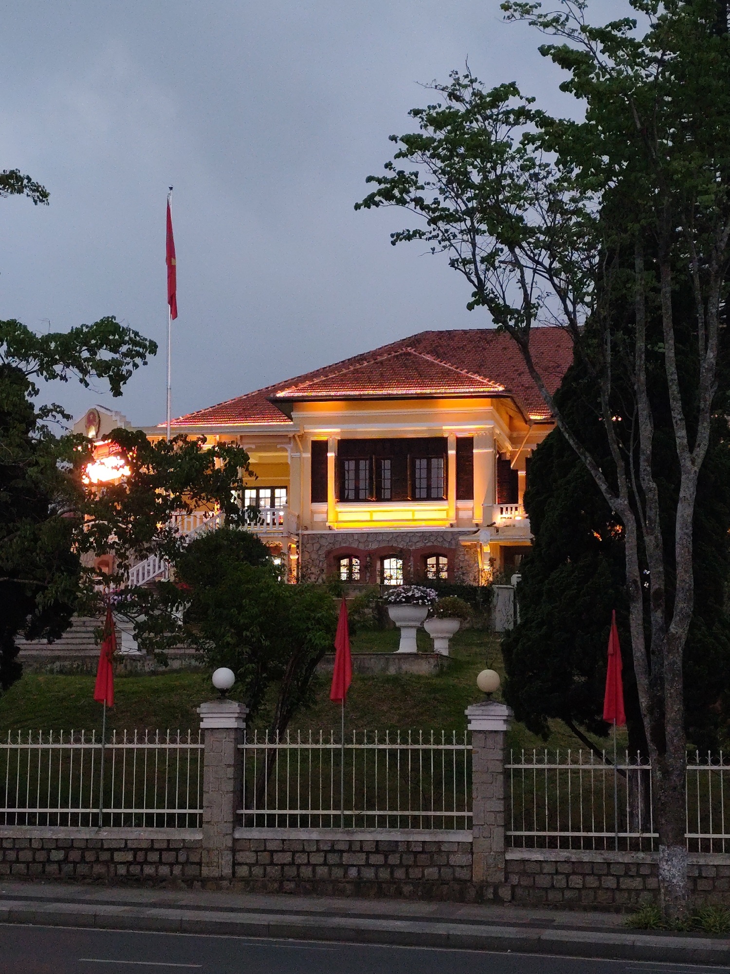 The image shows a stately yellow building illuminated by warm light in Da Lat, Vietnam. A red flag stands tall on a pole to the left of the building. The building is set on a small hill, accessible by stairs, with manicured green space. A white metal fence with stone pillars runs along the base of the hill, bordering a road. There are several red flags placed on the hill near the fence. The sky is overcast, suggesting dusk or dawn. Trees are visible framing the building.