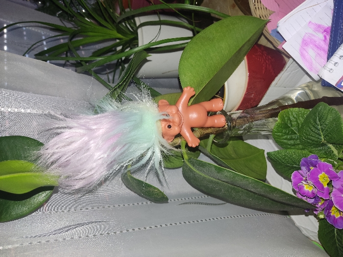 A vintage troll doll with pink and blue hair lies beside a glass bottle. The scene is decorated with green leaves, a purple flowering plant, and a white, sheer fabric. A potted plant, papers, and a basket are blurred in the background. The troll doll is small, with a tan body and its arms and legs outstretched.