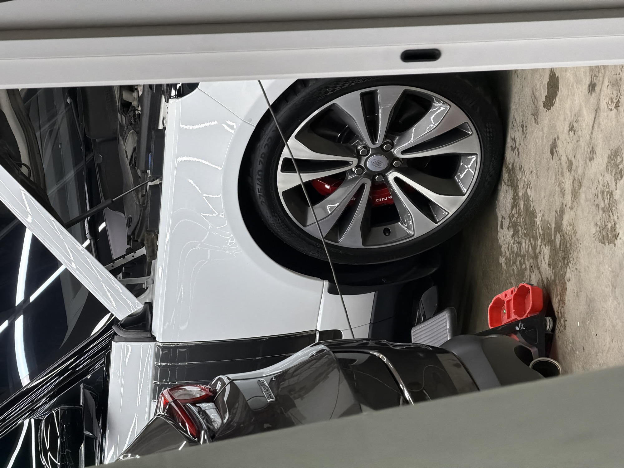 The image shows a white and gray Land Rover vehicle with its hood open, parked in an enclosed space, possibly a garage, in Krasnoyarsk, Russia. The vehicle's front passenger side wheel is visible with a multi-spoke silver and black rim and red brake calipers. A red and black object, which might be a tool container, sits on the floor next to the wheel. The environment appears clean and well-lit.
