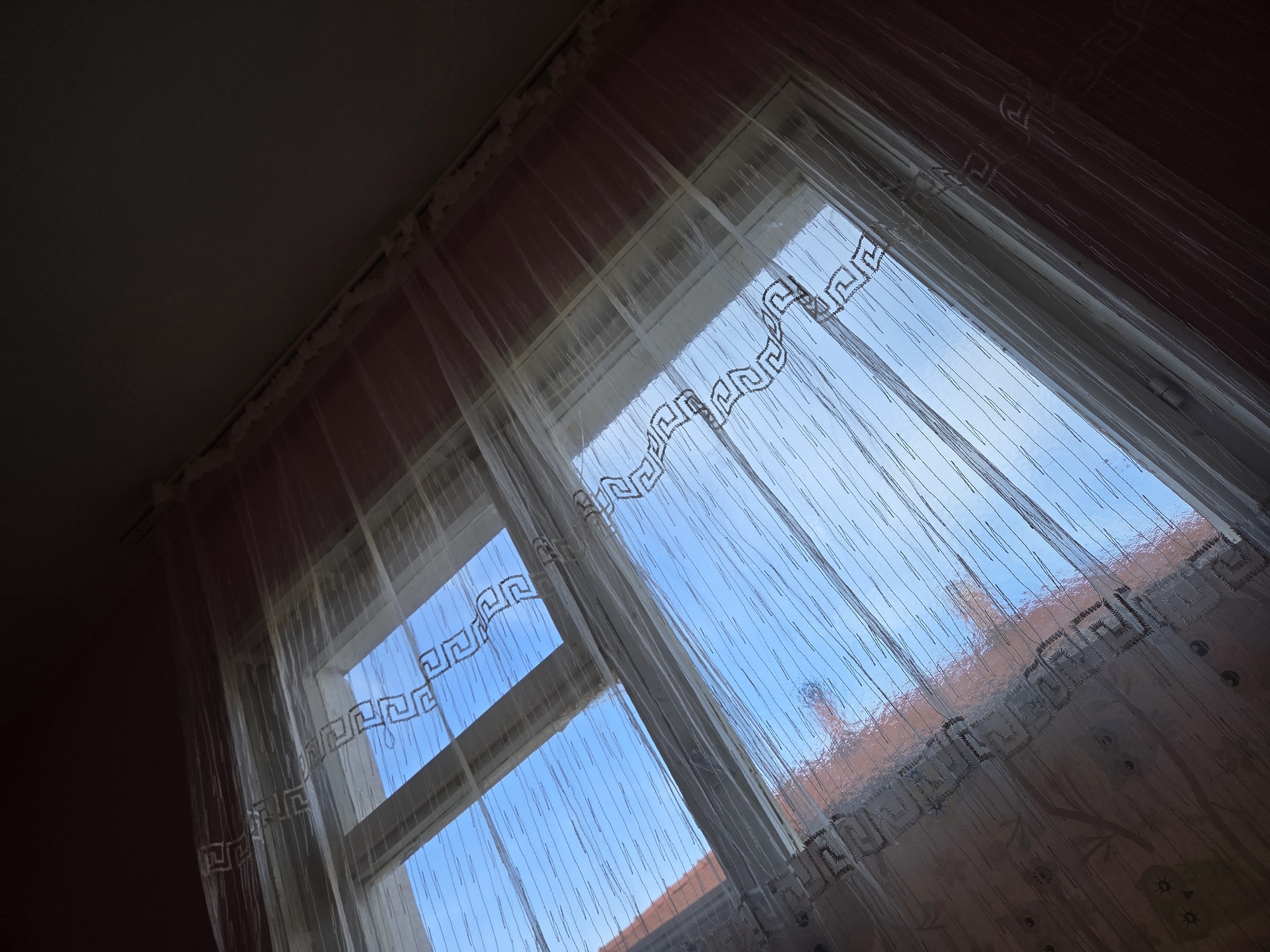 The image presents an indoor perspective looking out of a window, captured from an upward, oblique angle. A translucent, sheer white curtain, adorned with a repeating dark grey or black pattern resembling a Greek key or meander, hangs draped across the window.
Through the window and curtain, a vibrant, clear blue sky is visible, indicating daytime with sunny weather. A glimpse of a reddish-orange tiled roof, likely from an adjacent structure, can be seen in the lower right portion of the outdoor view. The window itself has a white frame, with vertical and horizontal divisions.
Indoors, the ceiling above the window is dark grey or black, while the surrounding walls are a reddish-pink hue. The interior appears relatively dim in contrast to the bright natural light filtering through the window and curtain. No people or specific activities are depicted in the scene, which offers a quiet, static view. No visible text is present. The setting is consistent with a residential environment in Krastevich, Bulgaria.