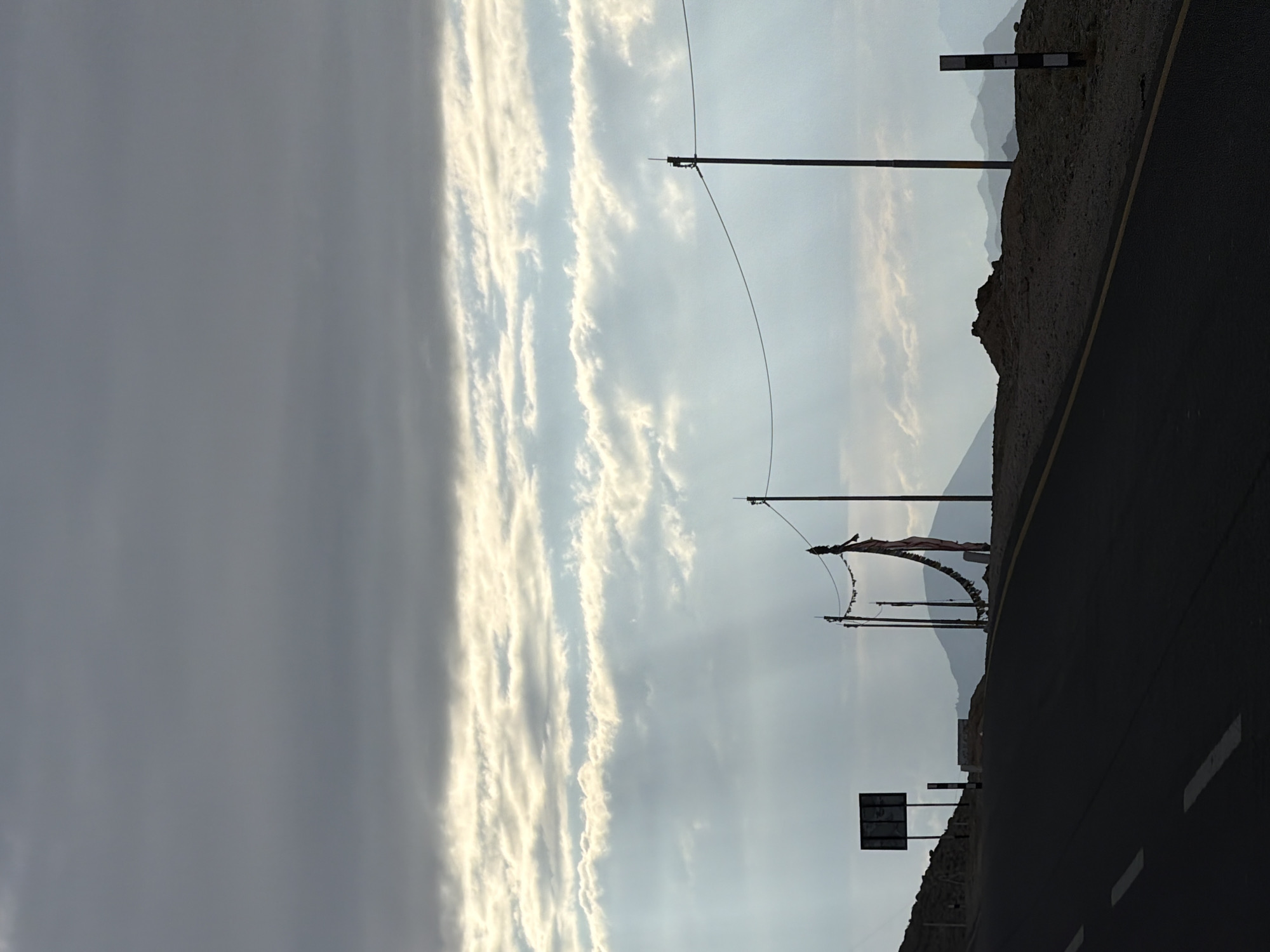 This image captures a desolate mountain road in Chumkar, India, under a dramatic, cloudy sky. The foreground is dominated by a dark asphalt road with white dashed lines. To the left, a hill rises, topped with a silhouette of a signpost. Further along the road, several utility poles stand, one strung with prayer flags that flutter in the wind. In the distance, a chain of muted mountain ranges stretches across the horizon, partially obscured by haze and the low-hanging clouds. The overall impression is one of solitude and vastness.