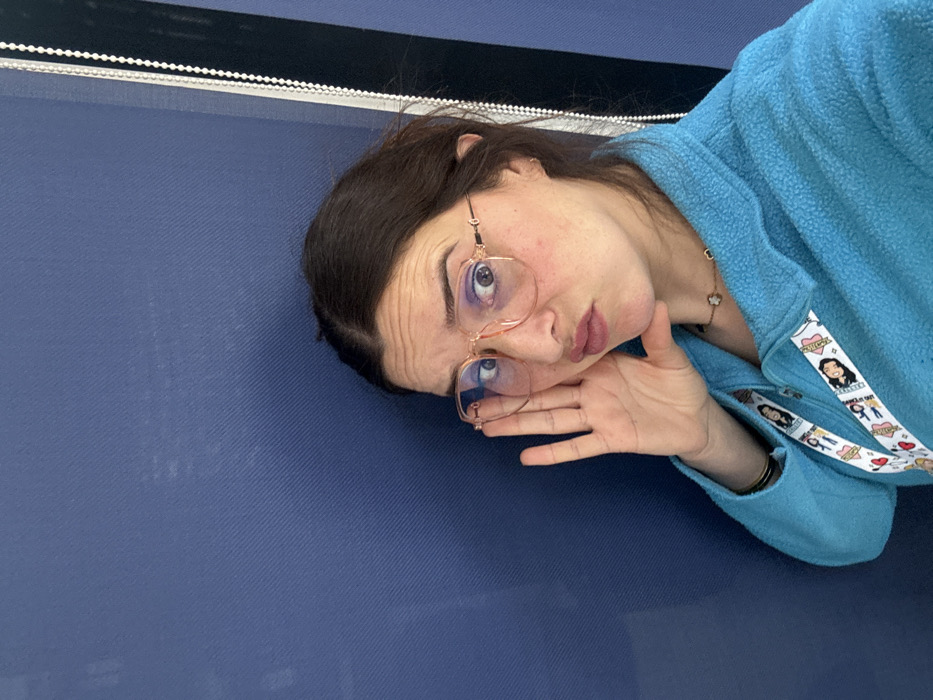 The image displays a single adult female in a headshot, positioned slightly off-center to the right against a plain dark blue background, likely an interior wall or partition with a subtle diagonal texture. To the far right, a vertical white beaded chain, consistent with a window blind or curtain mechanism, is visible.
The female has dark hair, wide-open eyes, and a furrowed brow. She is wearing large, light peach-framed, octagonal-shaped glasses. Her lips are pursed in a slight pout. Her right hand is raised with fingers spread, resting against the side of her face and jawline, creating a posed expression.
She is dressed in a light blue fleece jacket with a partially visible zipper. Around her neck, a delicate gold-toned necklace with a black four-leaf clover pendant is present. A white lanyard, featuring cartoon female characters and text, is worn around her neck, partially visible beneath her jacket collar. A thin, dark bracelet is visible on her left wrist.
There are no discernible visual elements within the image to specifically identify the location as Meknes, Morocco; the background is generic and provides no city-specific context.