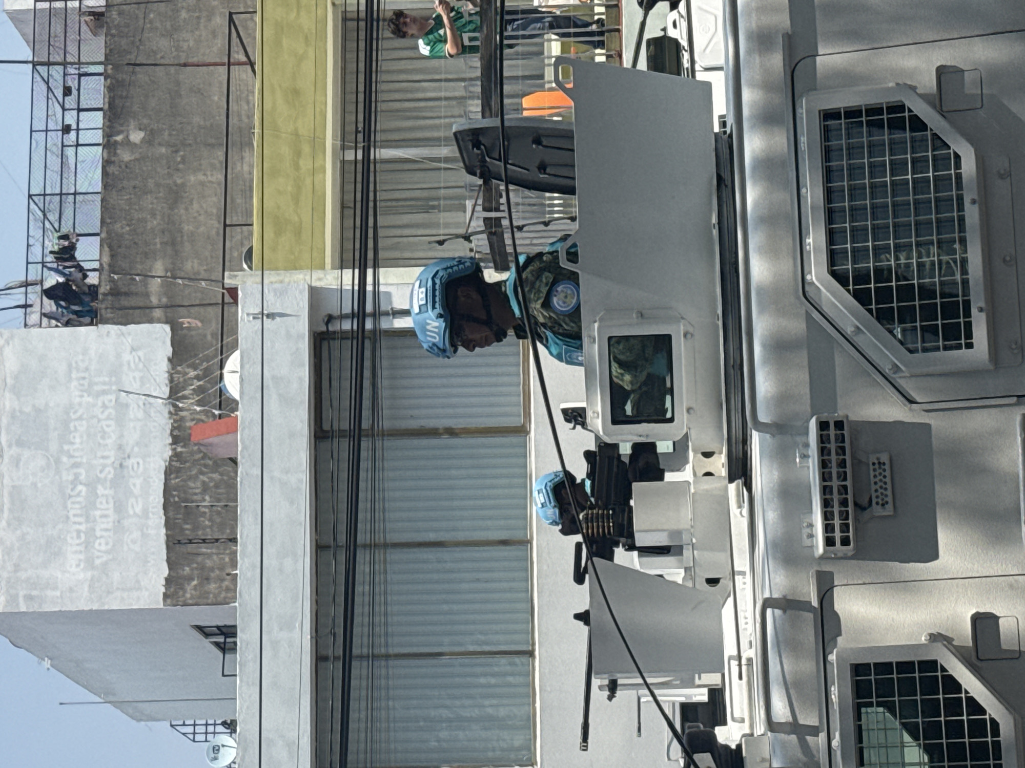 A soldier wearing a blue UN helmet is stationed inside a UN armored vehicle. The soldier is facing left in the image, with a gun and ammunition belt visible near him. Outside the vehicle, there are buildings and power lines, suggesting an urban setting. A person wearing a green and white shirt is visible on a balcony. The environment appears to be the city of Puebla, Mexico. A building with "Tenemos 5 ideas para vender Su casa!!!" is visible on the side.