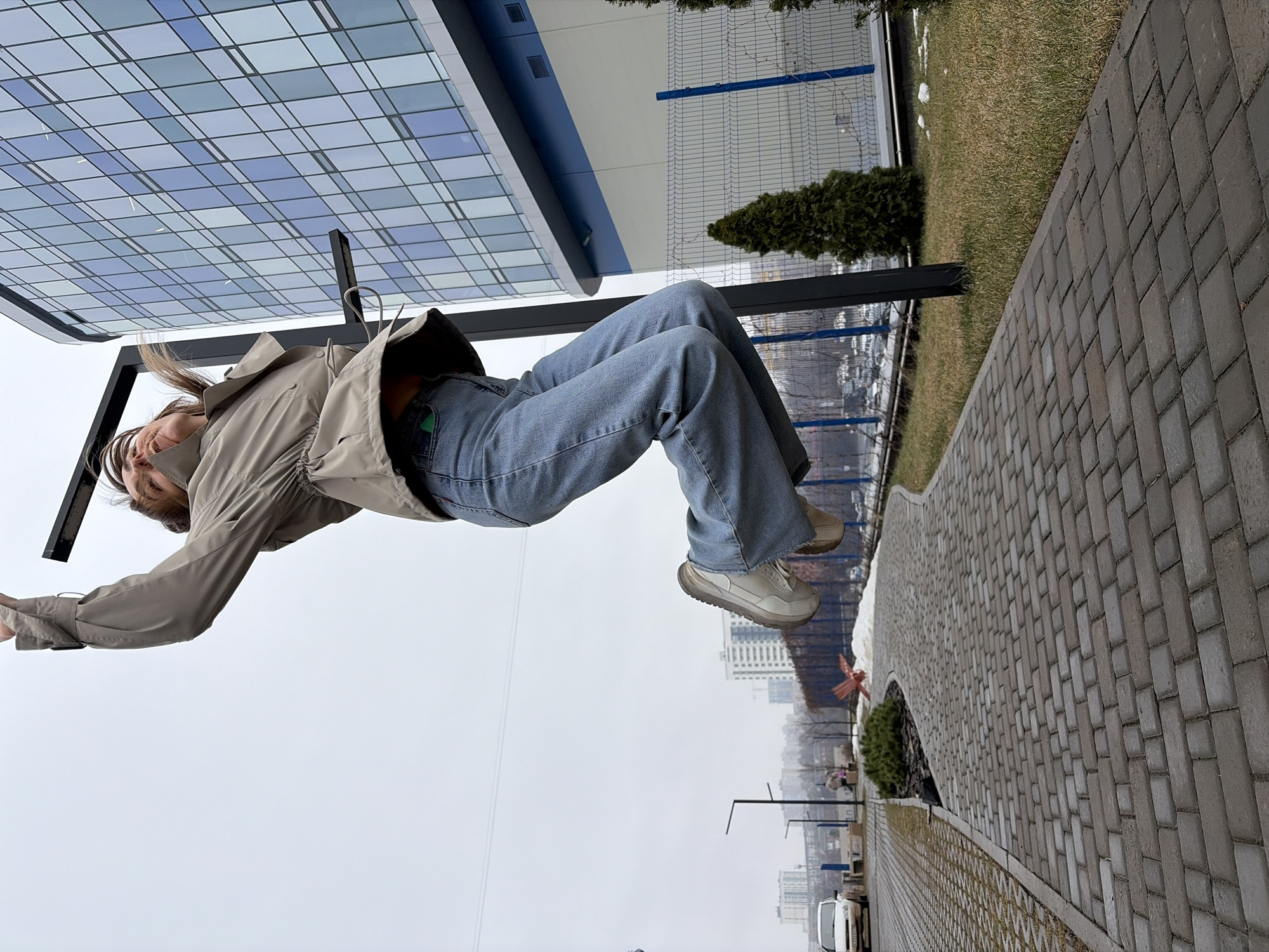 A woman is seated on a metal bench under a modern building with a blue and gray grid-like pattern. She wears a light-colored jacket, blue jeans, and light-colored sneakers. The image appears rotated, giving the impression she is suspended in mid-air. Behind her is a chain-link fence, grass, and a small tree. In the distance, buildings and city structures are visible. The photo shows a paved walkway composed of gray bricks. Based on visual cues, it's Izhevsk, Russia.