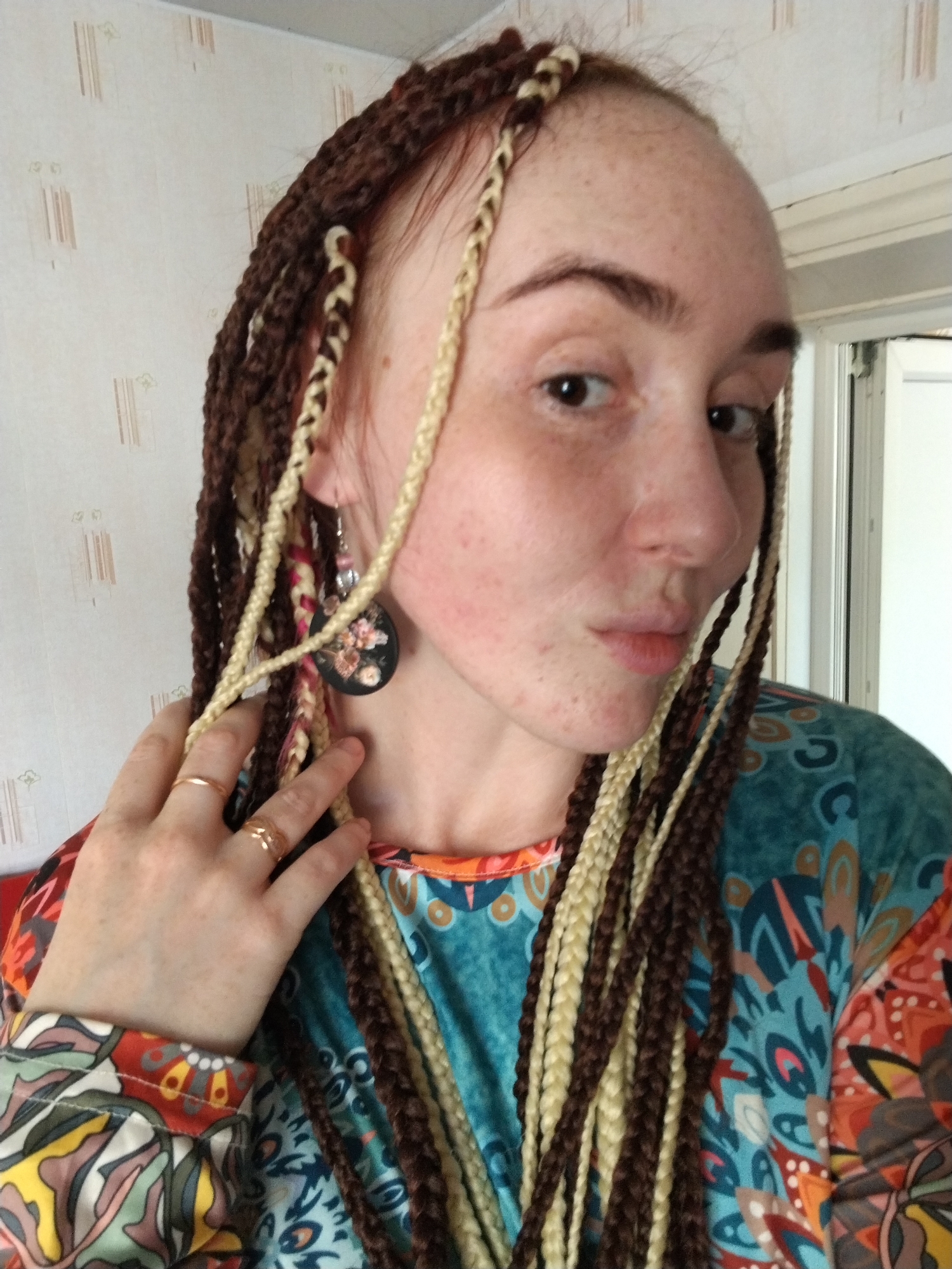 A fair-skinned person with brown and blonde braids is posing indoors. The person is wearing a colorful patterned top and earrings with a black floral design. Two gold rings are visible on their hand. The background includes a wall and a partially visible white door frame. There is no definitive visual information in the photo to identify the location as Яган, Russia.