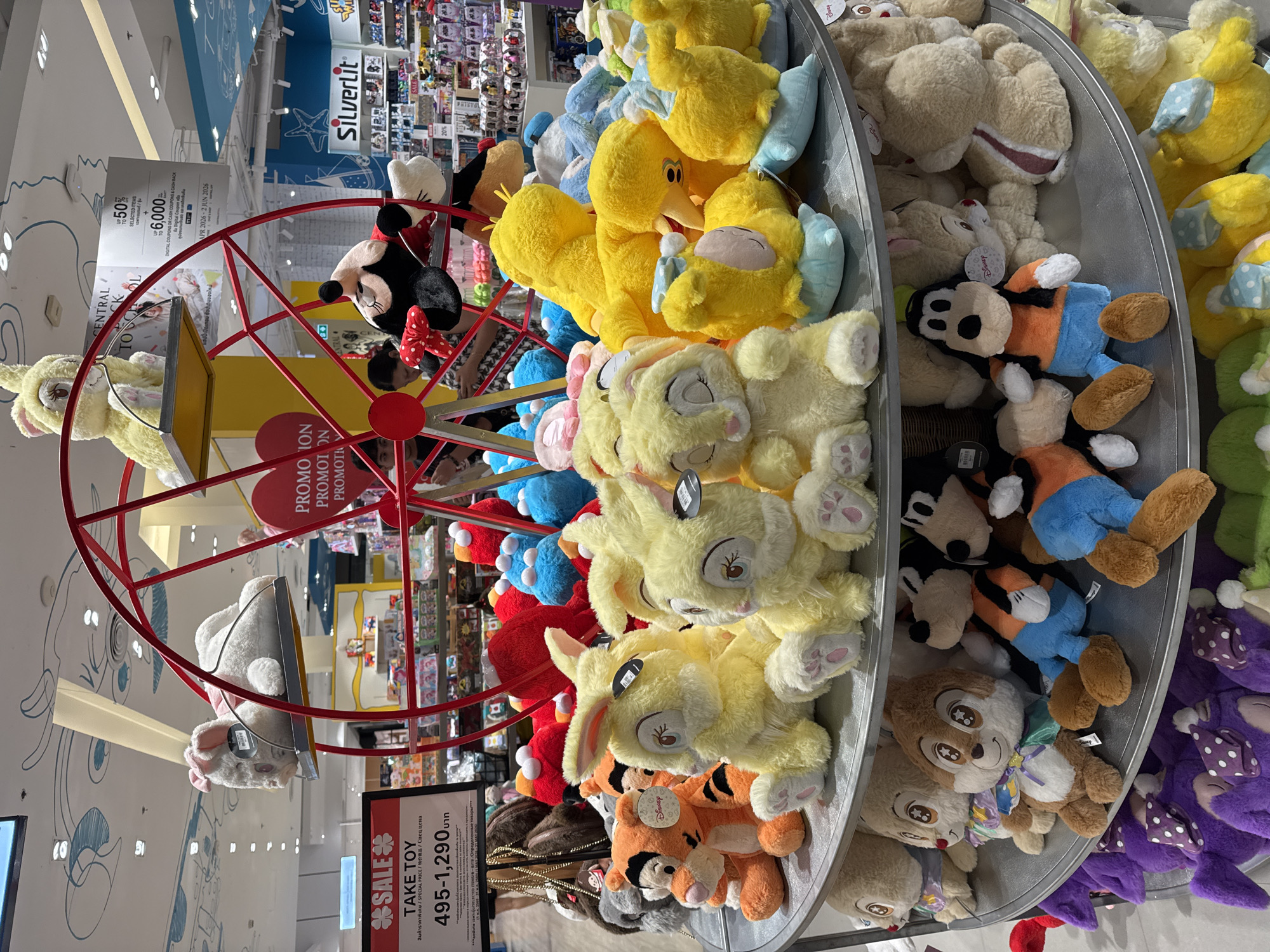 The image shows a display of stuffed animals in a store. The display is arranged like a Ferris wheel, with various plush toys in each section. There are Disney characters like Mickey Mouse, Big Bird, and Thumper. A sign indicates a sale on "TAKE TOY" items, priced between 495-1,290 baht. Other signs mention promotions and coupons. The location is Baan Chaweng, Thailand.