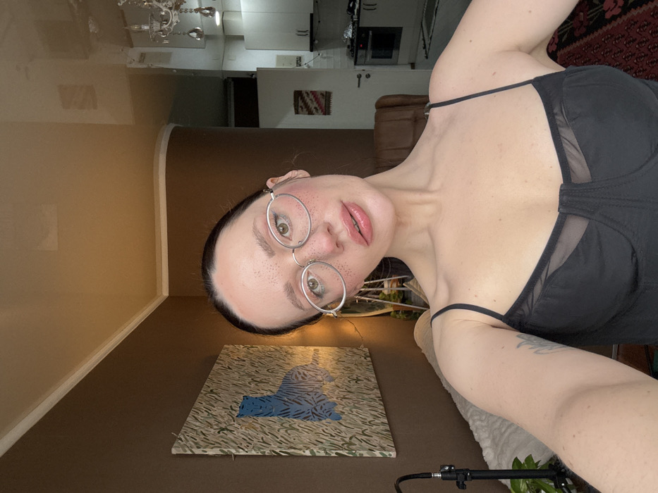 This indoor selfie photograph from Batumi, Georgia, features a female subject in the foreground, looking directly at the camera with her head tilted slightly right. She has dark hair, wears light-framed oval eyeglasses, and displays prominent freckles on her face. Her lips are parted and glossy. She is dressed in a black camisole with thin straps and mesh bust detail; a tattoo is visible on her left bicep. The background on the left consists of a dark brown wall adorned with a large square painting depicting a blue tiger or cat in green foliage. A black stand with a plant and a white textured item are partially visible below. The background on the right reveals an open-plan living or kitchen area with lighter walls. Visible elements include a crystal chandelier, a white kitchen with white cabinets and a built-in dark oven, and a small, framed geometric textile art piece. A brown upholstered armchair is partially visible, and a patterned rug is seen on the floor in the lower right. The ceiling is light beige with white crown molding.
