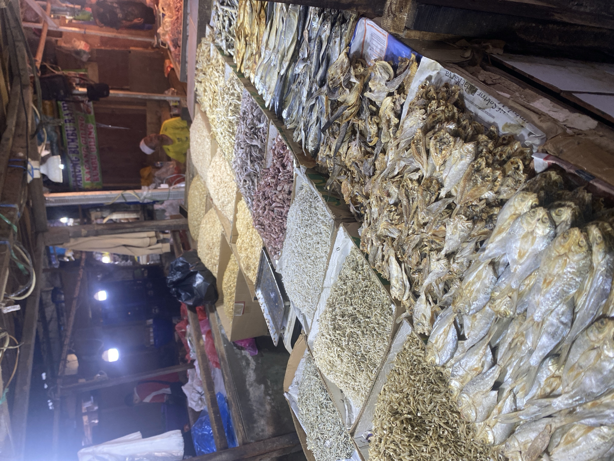In the city of Cikupa, Indonesia, a market stall displays a variety of dried goods, primarily fish and what appear to be grains or legumes. The stall is covered by a wooden structure with exposed wiring and lighting. Multiple cardboard boxes and trays are filled with these items. In the background, a person wearing a yellow shirt and a white cap is visible behind the stall. A sign with Indonesian text, "PARUNG BUMBU RAJE," is hanging above. The lighting is dim, with several bulbs illuminating the scene. The overall impression is that of a local marketplace with abundant dried produce for sale.