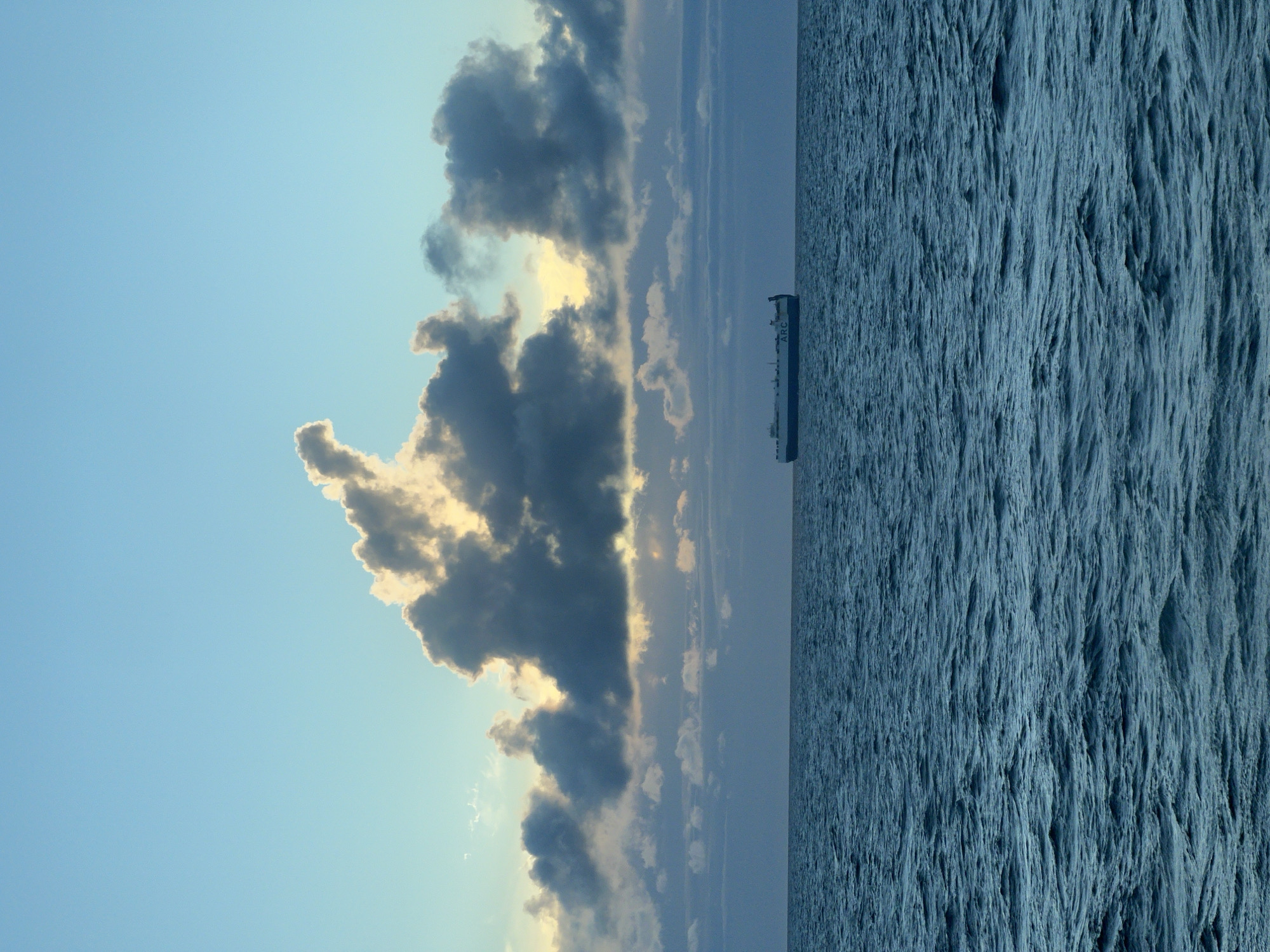 A large cargo ship, identified by the white text "ARC" visible on its side, sails across a deep blue ocean under a sky filled with dramatic, illuminated clouds. The clouds are dark and stormy in places, with bright sunlight piercing through some of them, creating a striking contrast. The sea exhibits moderate wave activity, indicating a dynamic environment. The horizon line is clearly defined, separating the vast expanse of the ocean from the cloudy sky. The image appears to be taken during either dawn or dusk due to the quality of the light.
