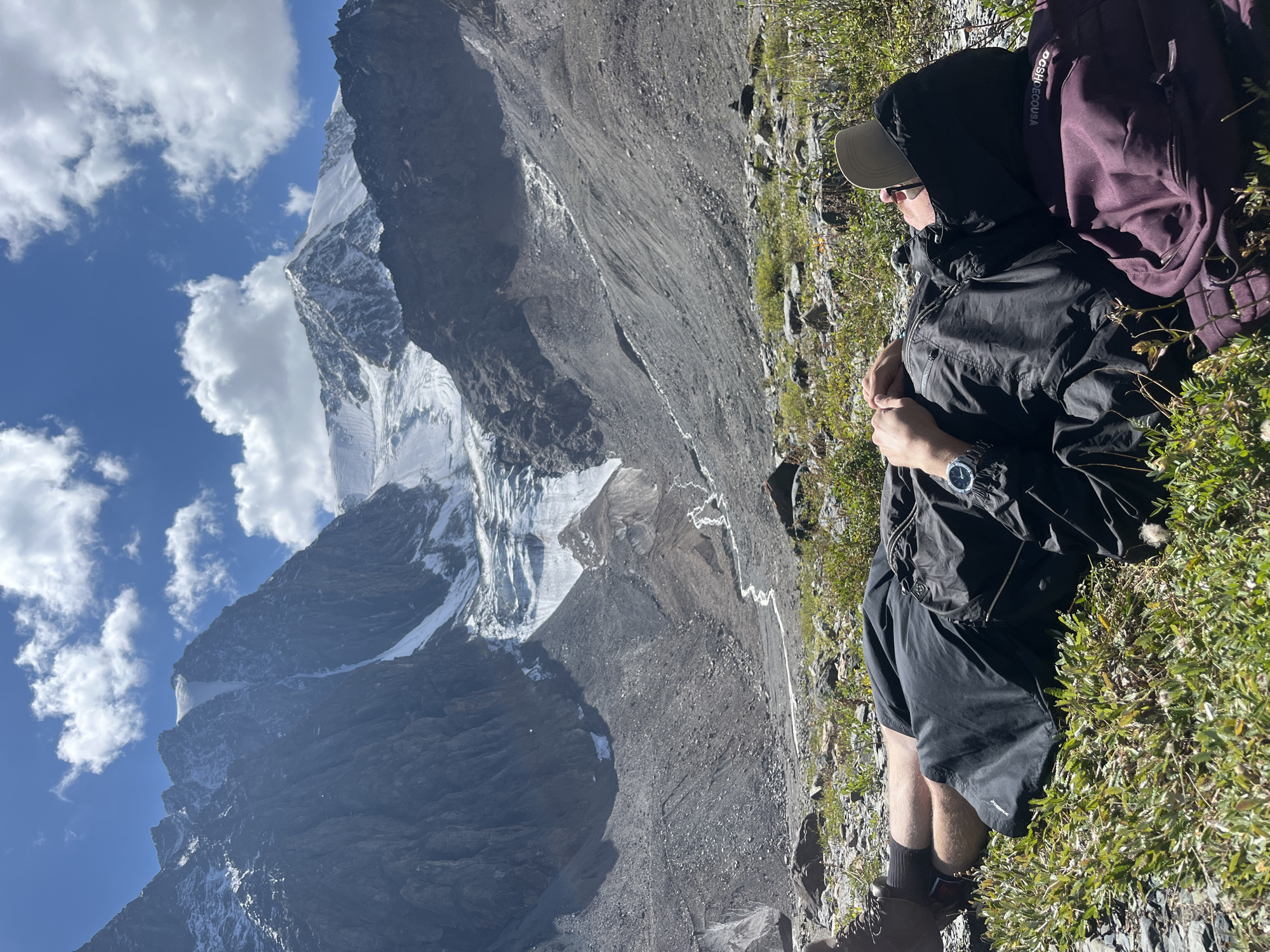 A person lies on their back in a grassy, rocky area with a large, snow-covered mountain range in the background. The person is wearing a black hooded jacket, black shorts, and brown hiking boots. A tan baseball cap is on their head, and they have sunglasses on. Their arms are crossed over their chest, and a burgundy backpack is to their right. The mountain range features steep, rocky slopes and a large glacier. The sky is blue with scattered white clouds. The location is identified as Unknown, Russia.