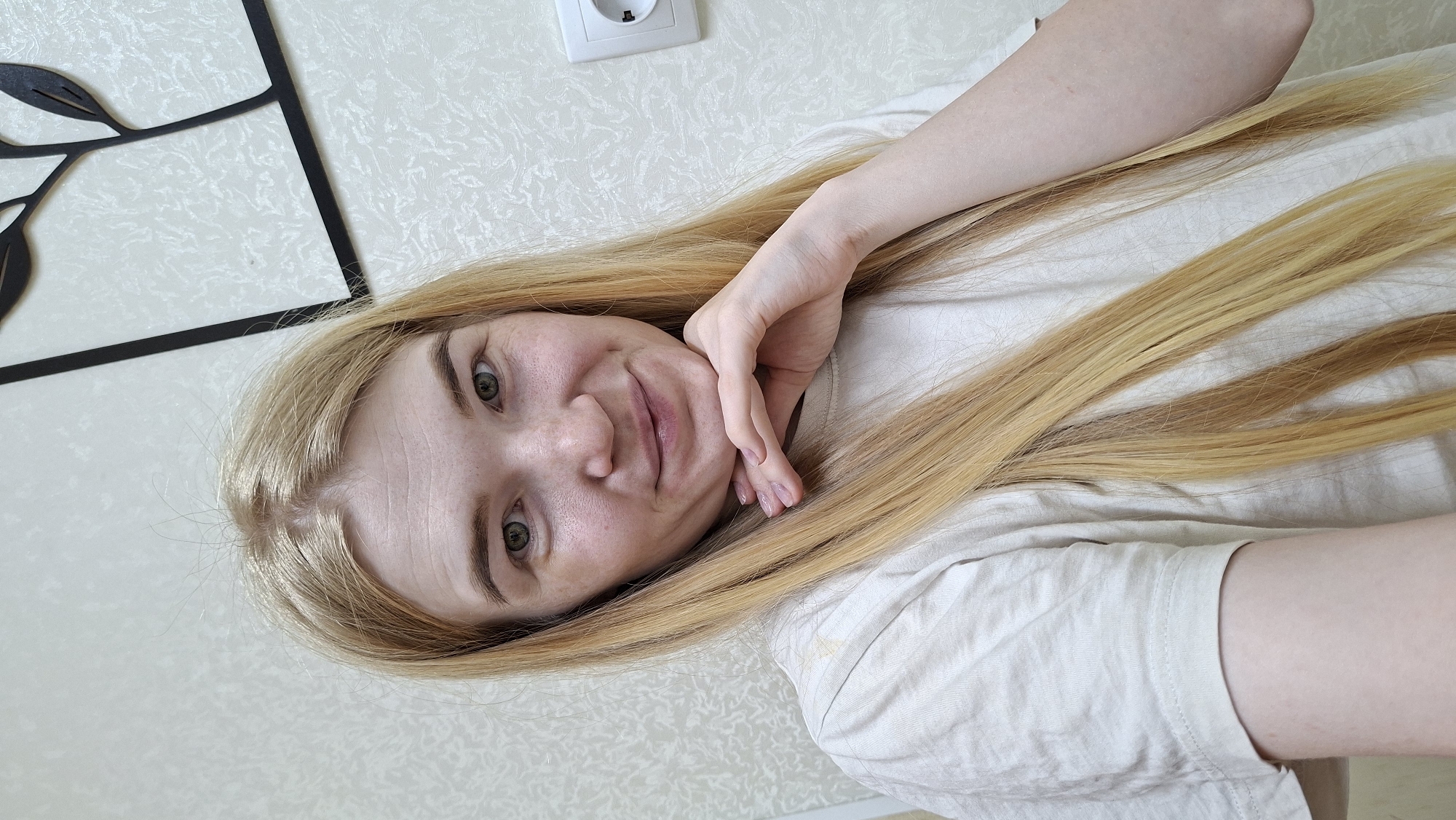 A woman with light blonde hair is lying down, her head at the bottom of the frame and her feet presumably towards the top. Her right hand is resting on her face. She has fair skin and is wearing a white T-shirt. Above her head, there is a white wall with a black metal decoration of leaves on the left and a white electrical outlet towards the top. The location is potentially in a residential setting in Большой Букор, Russia, although specific indicators of the city are not visible in the image.