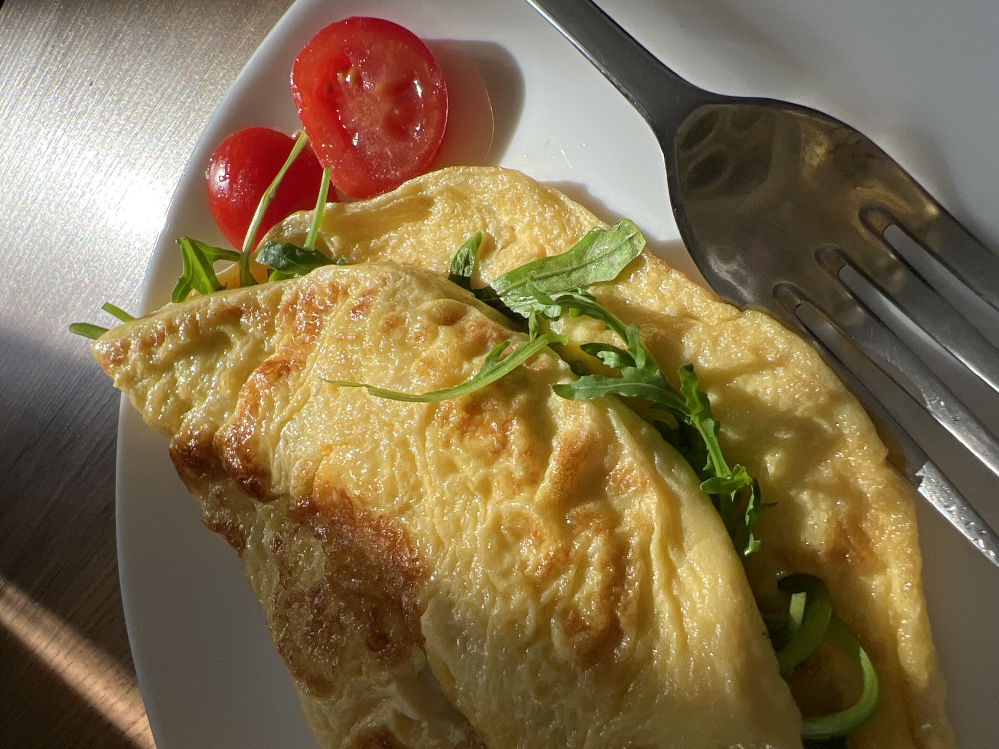 A plate contains a folded omelet garnished with arugula and sliced cherry tomatoes, with a fork placed beside it. The plate is white and reflects light, suggesting a shiny or glossy surface. The omelet has a browned texture in places, indicating it has been cooked. The arugula is green and fresh, providing a contrasting color to the yellow omelet. The cherry tomatoes are red and sliced in half. A fork is set on the right side of the omelet, appearing made of stainless steel. The scene is brightly lit, casting shadows that suggest it is daytime. There is no information about the location being in Kazan, Russia.