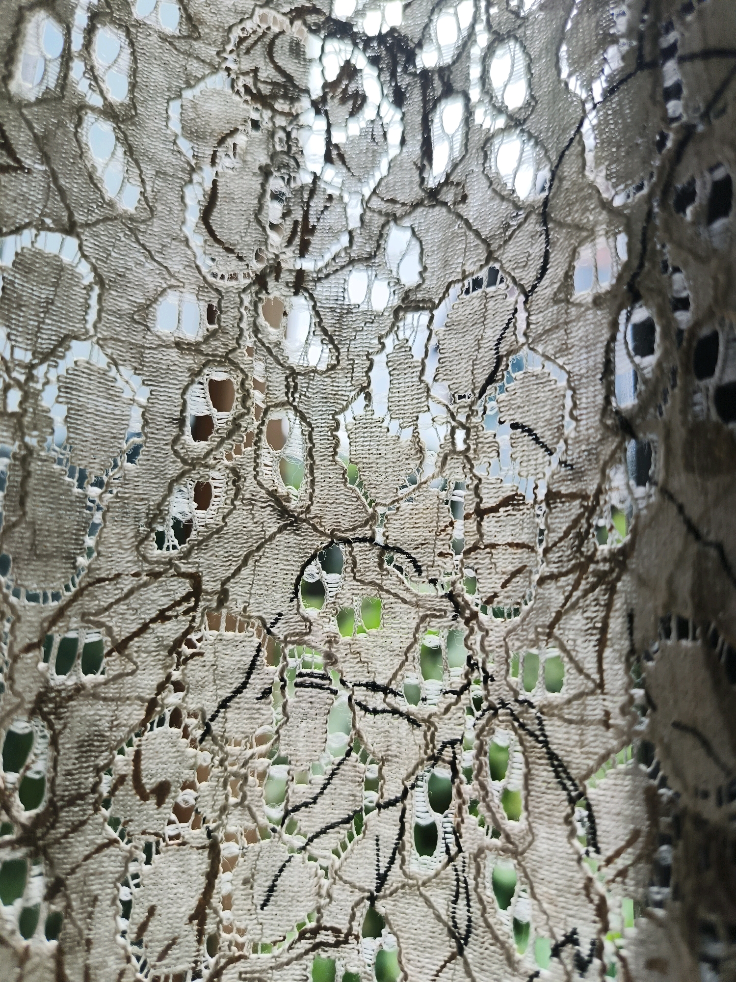 A close-up shot of a beige-colored lace curtain with an intricate pattern of holes and dark brown embroidered lines. The curtain is translucent, allowing light to filter through and reveal a blurred background of green foliage and some light blue sky. The texture of the lace is rough and uneven, with some areas appearing frayed. The image is taken in Aizawl, India.