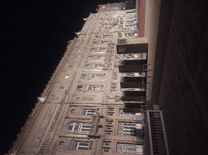 A low-angle shot captures an ornate, tall building at night. The structure features numerous windows of varying sizes, some arched. Intricate decorative moldings and sculptural details adorn the facade. The building's exterior is illuminated, contrasting against the dark night sky. Modern metal structures, possibly bike racks, line the lower portion of the building, obscuring some of the ground floor windows. A dimly lit wall stretches to the right, creating a strong contrast.