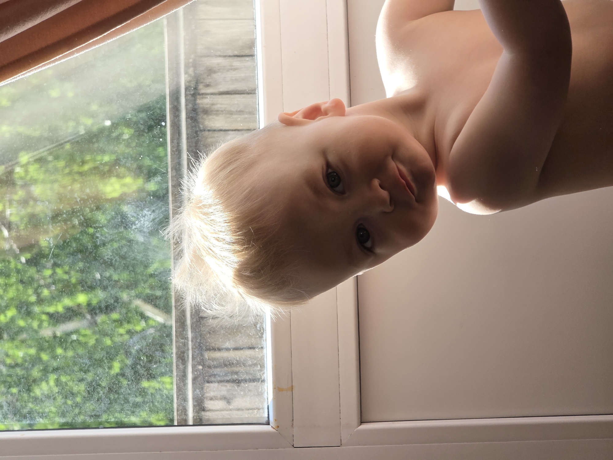A baby with light blond hair and fair skin is tilted sideways, positioned near a window. The window is visible on the left, with a view of green foliage outside. The baby's expression is somewhat inquisitive. The room seems brightly lit, as evidenced by the light on the baby and the window frame. A curtain can be seen at the top of the image. There is no indication of the location being Chaikovsky, Russia.
