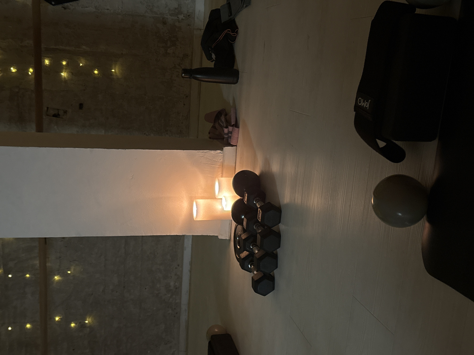 This is a dimly lit image of an indoor space, likely a fitness studio or home gym in Tel-Aviv, Israel, as suggested by the context. The floor is light-colored wood, and the walls appear to be concrete with some decorative elements. The scene is illuminated by soft, warm light from several pillar candles placed near a white pillar in the center of the frame.
Dominating the foreground are several black hexagonal dumbbells arranged in a cluster on the floor. To their right, a black water bottle stands upright. Behind the dumbbells and to the right of the pillar, a pair of pink exercise bands are neatly folded. Further back, on the floor, is a black yoga mat with a small, light-colored sphere, possibly a small exercise ball, resting on it. To the far right, a black bag with the brand "JOMO" is visible. The background walls are textured and adorned with strings of small, warm-toned fairy lights. The overall atmosphere is serene and inviting, suggesting a space set up for exercise or relaxation. There are no people visible in the image.