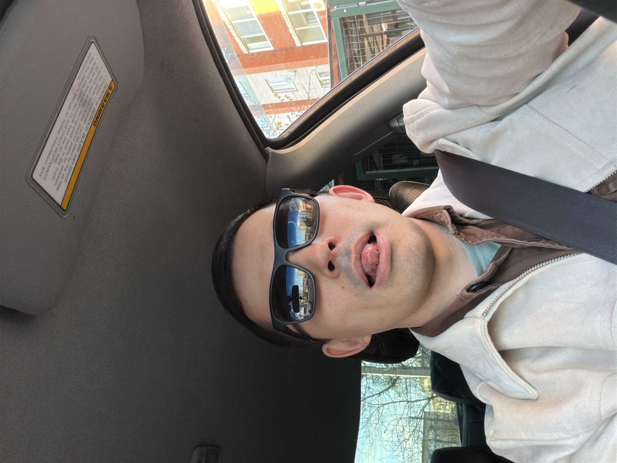 The image features a close-up, upward-angled view of a person, a male with dark hair, seated in a car, likely in the passenger seat. He is wearing dark sunglasses and a light-colored hooded jacket over a brown and light blue top, with a seatbelt secured across his chest. The subject is looking upwards and outwards, with his tongue playfully extended from his mouth.
The sunglasses reflect a clear image of the urban environment outside, including buildings and parts of the car's interior. Above, the car's sunroof is open, revealing multistory brick buildings with windows and a bright, clear sky, along with some bare tree branches, suggesting daytime with good weather. The car's gray interior headliner is visible, as is a sun visor with a prominent warning label.
The text on the warning label reads: "A WARNING. DEATH or SERIOUS INJURY can occur. Children 12 and under can be killed by the air bag. The BACK SEAT is the SAFEST place for children. NEVER put a rear-facing child seat in the front. Sit as far back as possible from the air bag. ALWAYS use SEAT BELTS and CHILD RESTRAINTS. See the Owner's Manual for further information and explanations."
The scene is set inside a vehicle within an urban environment, identified as Izhevsk, Russia. The bright light suggests it is daytime. The person's expression conveys a casual and playful mood.