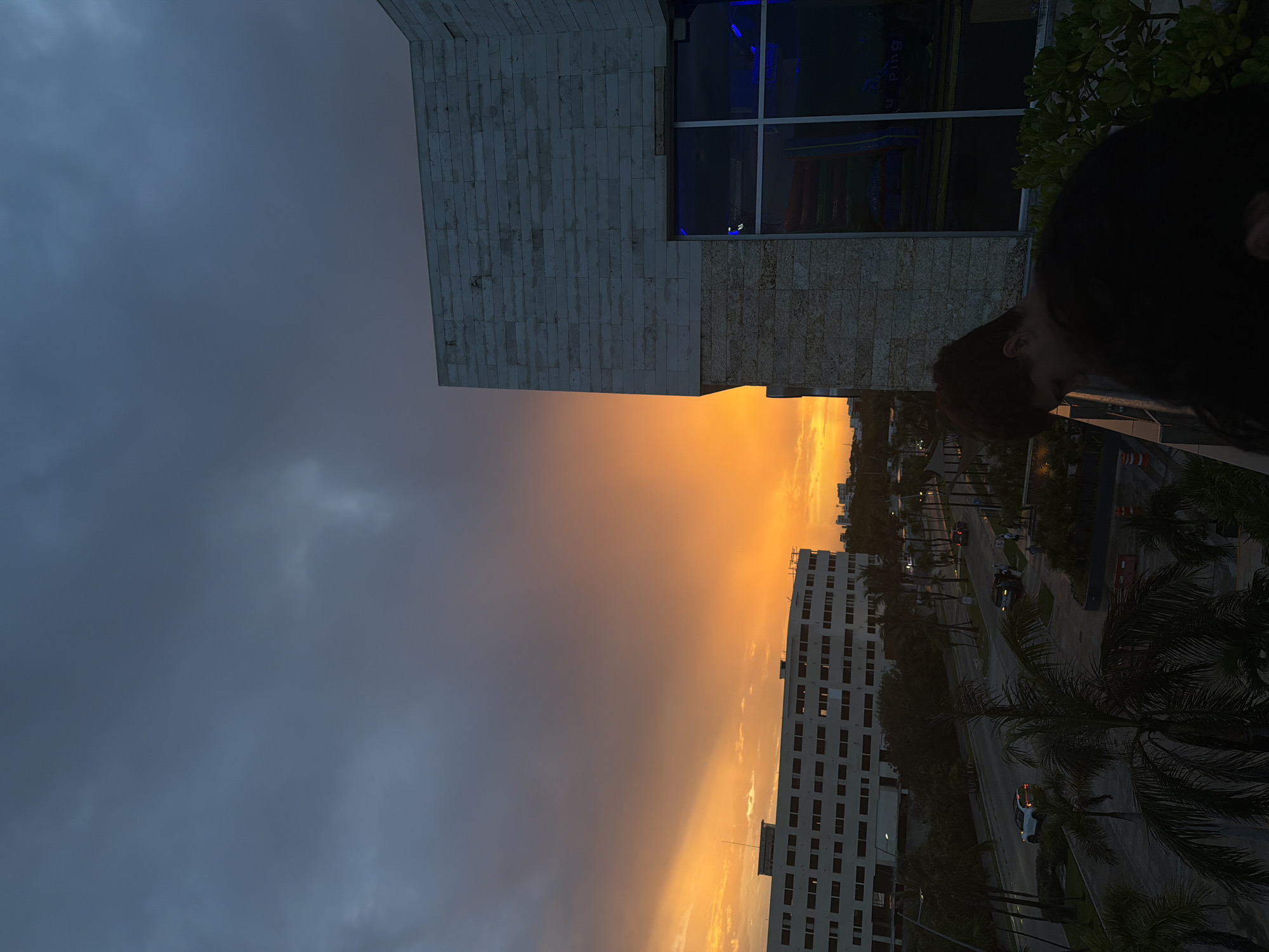 The photograph captures a dramatic sunset over Cancún, Mexico. The sky is ablaze with vibrant orange and yellow hues, with dark, ominous clouds gathering above. In the foreground, a young person in a black shirt is partially visible, leaning over a railing, seemingly looking down at the scene.
To the left of the image, a multi-story building with numerous windows is illuminated by the sunset's glow. It appears to be an office building or hotel, with palm trees and other vegetation lining the street below. Cars are visible on the road, suggesting urban activity.
To the right, a modern building with a stone facade features large windows with a blue interior light. The overall impression is one of a city preparing for dusk, with the natural spectacle of the sunset dominating the visual landscape. The image seems to have been taken from an elevated vantage point, possibly a balcony or rooftop, overlooking the cityscape.