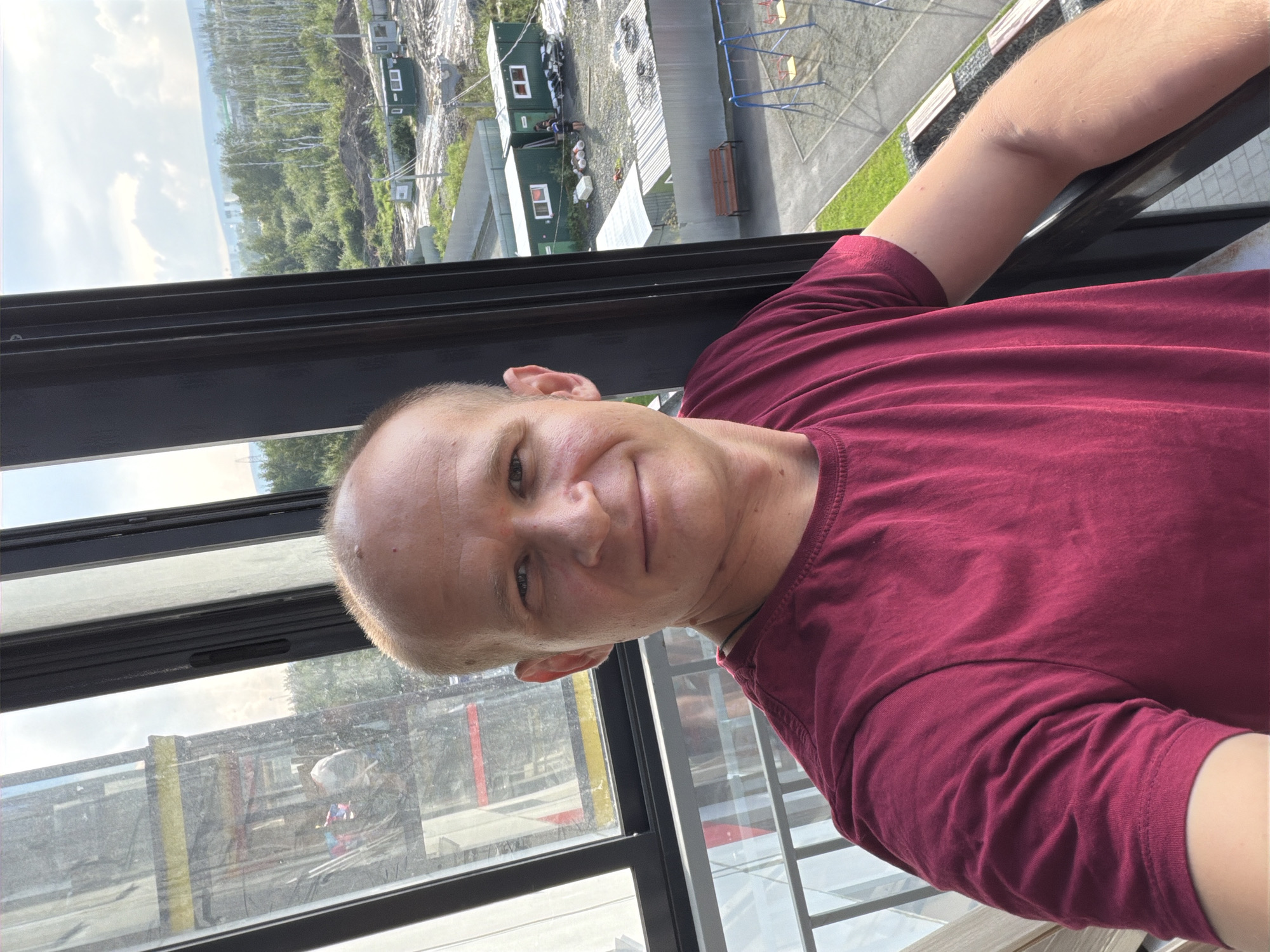 An outdoor selfie is taken from a high vantage point, likely an apartment balcony in Yekaterinburg, Russia. The foreground features a man with a shaved head and a slight smile, wearing a maroon t-shirt. He is positioned in front of a dark-framed window, which looks out onto a construction site and a residential area.
In the background, a large construction elevator is visible, with workers and materials present. Beyond that, the scene transitions to a developing neighborhood with several green portable buildings, likely temporary construction offices or worker accommodations. Further into the distance, a playground with swings can be seen, and beyond that, a landscape of trees and hills under a partly cloudy sky. The overall impression is of a modern urban development in progress, viewed from a residential building.