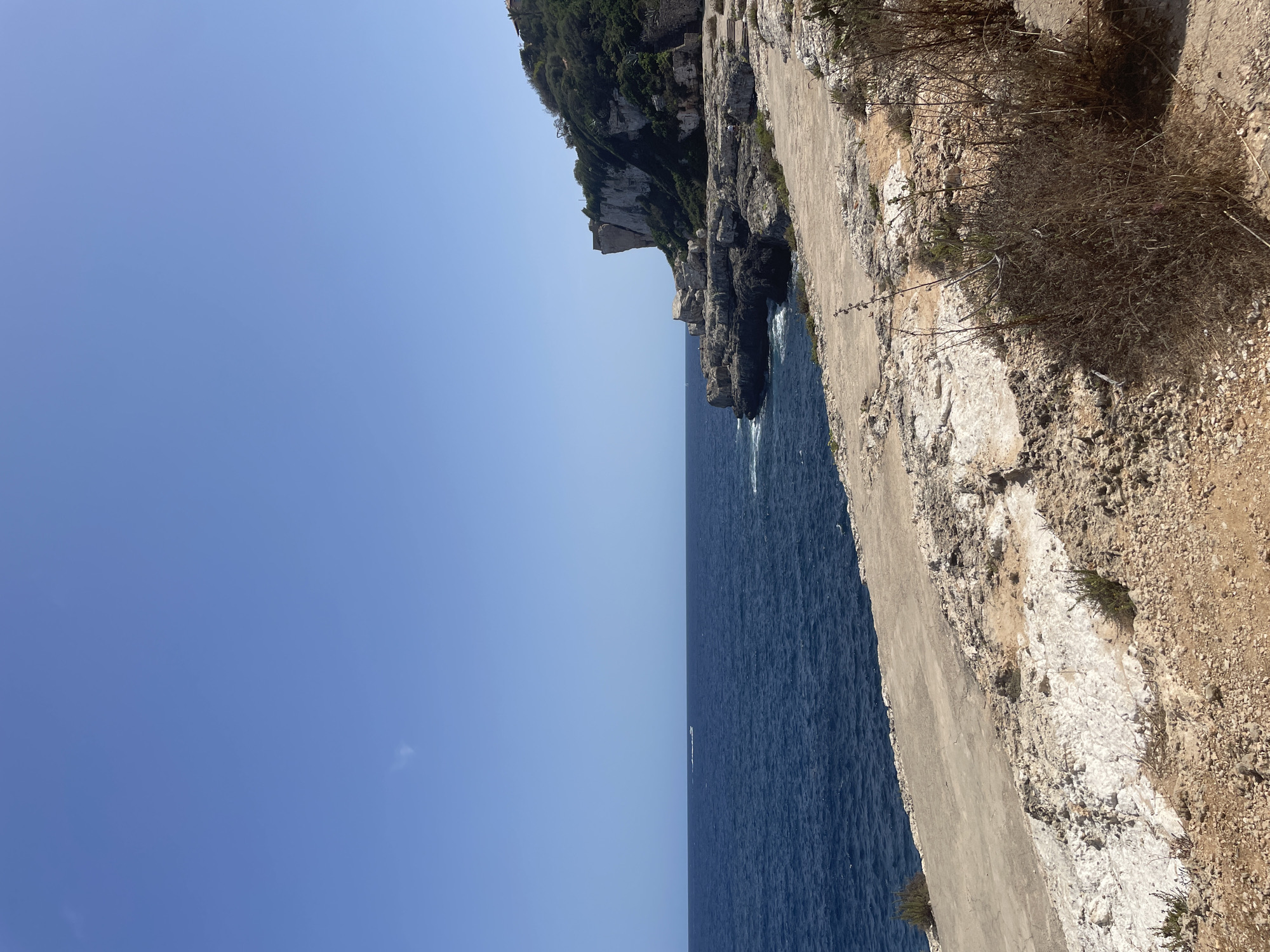 The image depicts a coastal landscape in the municipality of Santanyí, Spain, likely near the cliffs of Cala Llombards or a similar area. The foreground is a rocky, sun-baked terrain with patches of dry vegetation and a concrete path or paved area running along the edge. This path appears weathered and cracked in places.
Beyond the foreground, the deep blue Mediterranean Sea stretches to the horizon under a clear, bright blue sky. Gentle waves are visible near the shore, where they meet a rocky coastline. In the middle ground, a dramatic cliff face rises from the sea, covered in lush green vegetation on its upper slopes and showing exposed white rock in places. A small, weathered stone structure or ruin is visible on the cliff edge.
In the distance, a faint white boat is seen on the water. The overall scene is bathed in bright sunlight, suggesting a warm, clear day. There are no discernible people clearly visible, though some very small figures may be present on the rocky outcrops, too small to identify. The environment is natural and rugged, characteristic of the Mediterranean coastline.