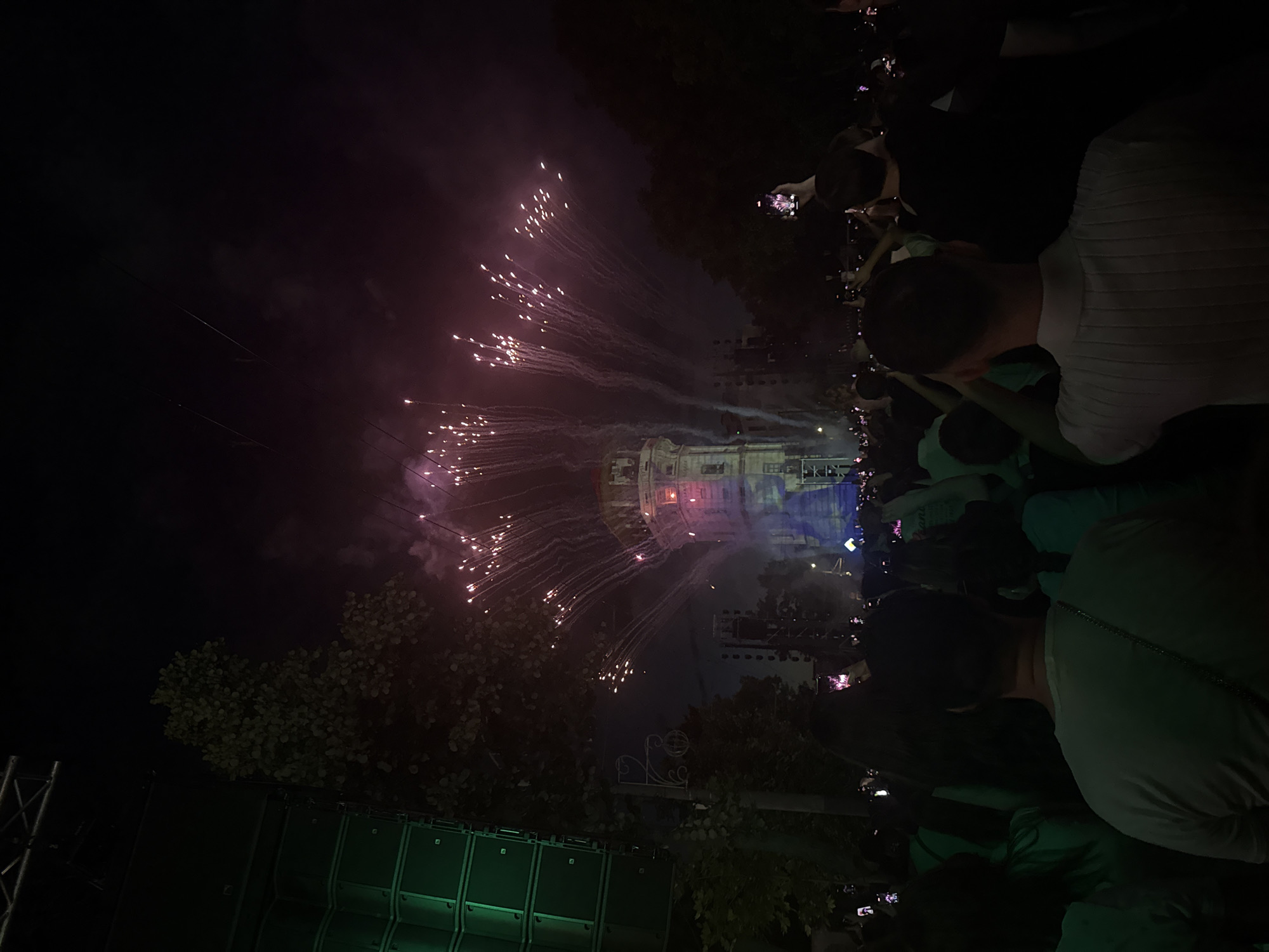 The image shows a nighttime outdoor event, likely a concert or festival, with a crowd of people in the foreground. Fireworks are exploding in the sky above a building, which appears to be a tower or a tall structure with architectural details. The building is illuminated with colorful lights, including purple and blue. To the left of the frame, a large green structure, possibly a speaker or stage equipment, is visible. The crowd is facing the building and the fireworks, and many people are holding up their phones to record the event. The overall atmosphere is festive and energetic. The presence of the National Opera and Ballet Theatre "Maria Bieșu" which is a prominent landmark in Chișinău, Moldova, and known for hosting events and being illuminated during special occasions, suggests that this scene might be taking place near this location in Chișinău.