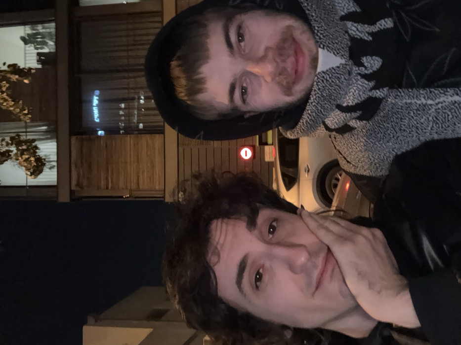 Two men pose closely together in an outdoor nighttime shot. The upper figure wears a hooded sweatshirt with patterned fabric, smiling slightly. The lower figure, sporting longer dark hair, rests his hand on his cheek. A building with windows and blinds is visible behind them, along with a white vehicle and a circular red sign. The lighting suggests an urban environment with streetlights nearby.