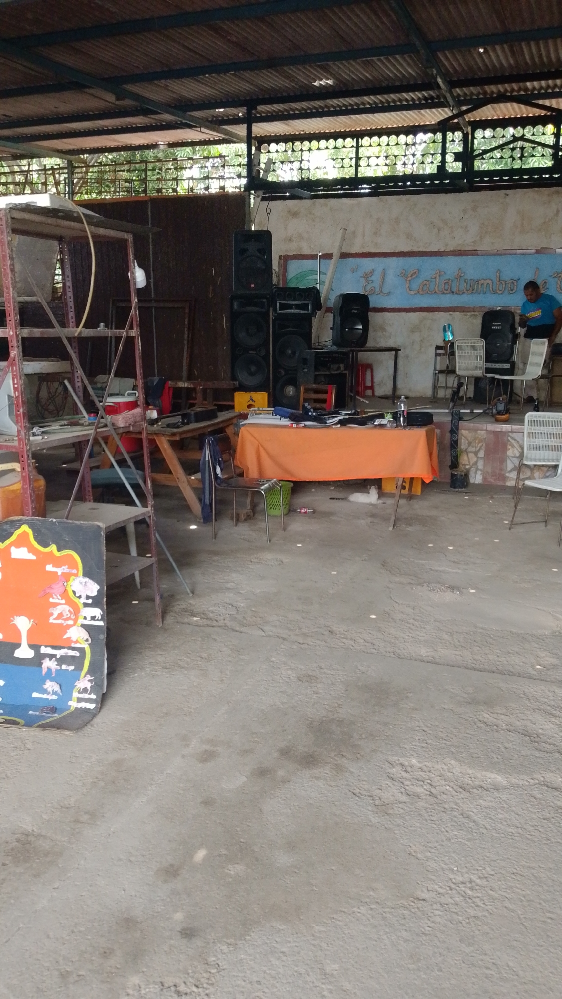 The image shows a large, covered open-air space, possibly a social or recreational area, in Tinaquillo, Venezuela. The space is utilitarian in appearance with a concrete floor and a corrugated metal roof supported by metal beams. There is a raised platform at the back with a painted mural bearing the name "El Catatumbo". A man wearing a blue shirt is partially visible on the platform. Several speakers are set up on the left side of the platform. An orange tablecloth covers a table in the middle of the area. There are chairs and other objects scattered around. A metal scaffolding structure stands to the left, along with a painted sign leaning against it. The overall impression is of a somewhat cluttered and unfinished space.