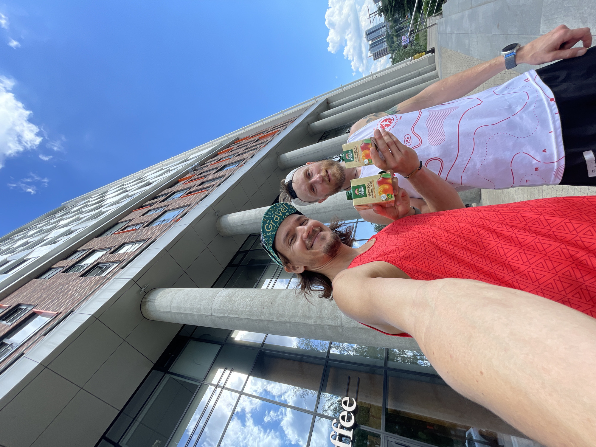 Two men are standing in front of a modern, multi-story building with large glass windows and columns. The building appears to be made of brick and has a grey facade. The sky is clear blue with some clouds.
The man on the left is wearing a red tank top and a green bandana with a pattern. He has long brown hair and is smiling at the camera. He is holding his arm out, as if taking a selfie.
The man on the right is wearing a white tank top with a red and white graphic design and black shorts. He has short blonde hair and a beard. He is holding two small cartons of juice. The cartons have an image of oranges and text in a foreign language. He is also looking at the camera.
There is a sidewalk and some stairs in the foreground. In the background, there are more buildings and trees. The overall scene suggests a casual outing or event in an urban setting.