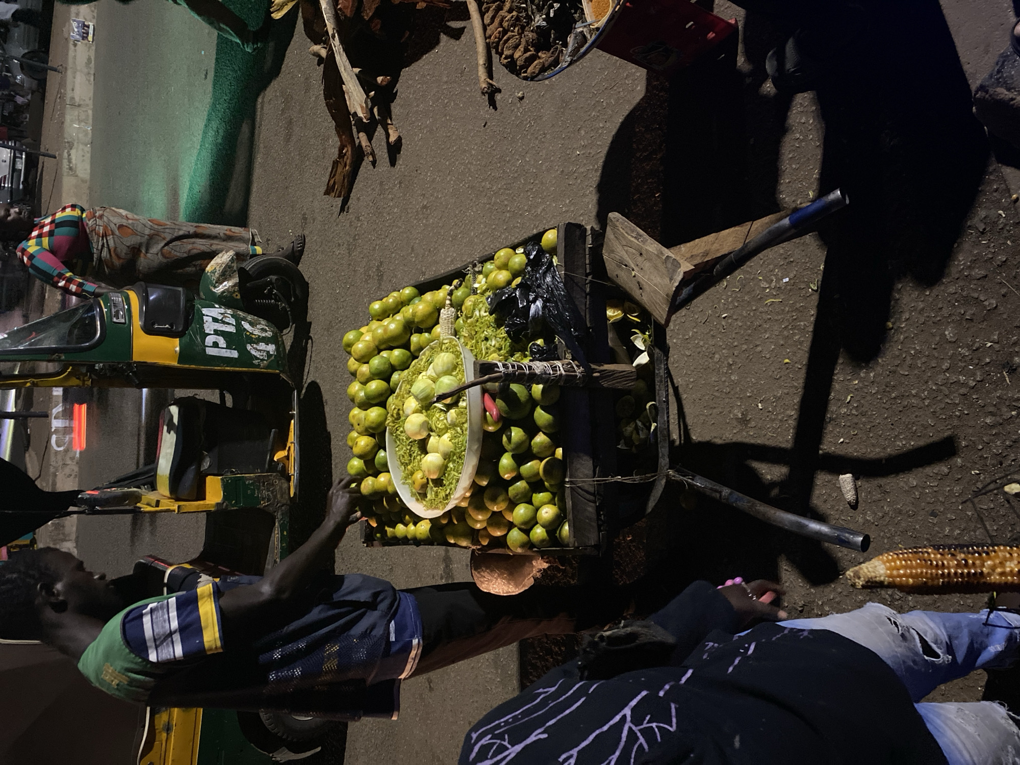 The scene is a street in Bukuru, Nigeria, likely at dusk or night. The image is from an overhead, slightly angled perspective. A three-wheeled auto-rickshaw is parked on the left, with a person wearing colorful clothes lying down, seemingly resting on its front. Beside the auto-rickshaw, a man wearing a blue and green shirt stands near a wooden cart piled with green fruits, possibly citrus like oranges or tangerines. The cart also contains a white bowl or container filled with sliced fruits. To the right of the cart, there's a person sitting, wearing ripped blue jeans and a dark hoodie. A few pieces of roasted corn lie on the ground near the sitting person. Further up the frame, there are dry branches and other objects. The ground surface is asphalt. Shadows suggest an external light source. The scene implies street vending and possible leisure activities in the evening.