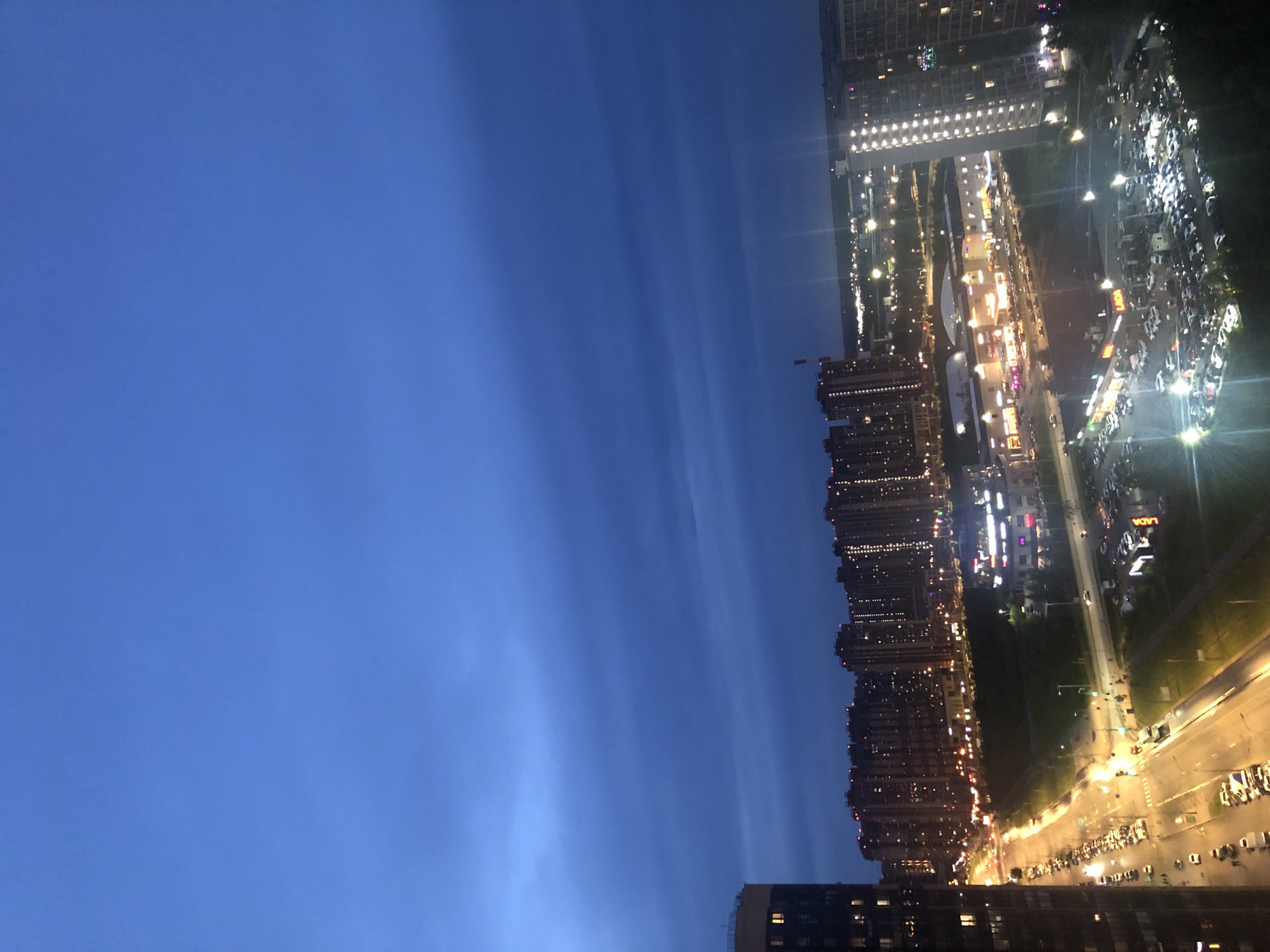This is a vertical, high-angle photograph of a city skyline at dusk. The sky is a deep blue with faint wispy clouds. On the right side of the frame, numerous tall apartment buildings are illuminated with countless windows glowing yellow. Below the buildings, a wide street is visible, lined with parked cars and streetlights casting a warm glow. The street appears to be a busy thoroughfare, with visible traffic and pedestrian activity suggested by the lights. In the distance, more city lights stretch out, creating a vast urban landscape. The overall mood is one of calm and transition, with the city coming alive as daylight fades. The time of day is clearly dusk, with the sky transitioning from day to night. No people are discernible in the image. There is no discernible text in the image.