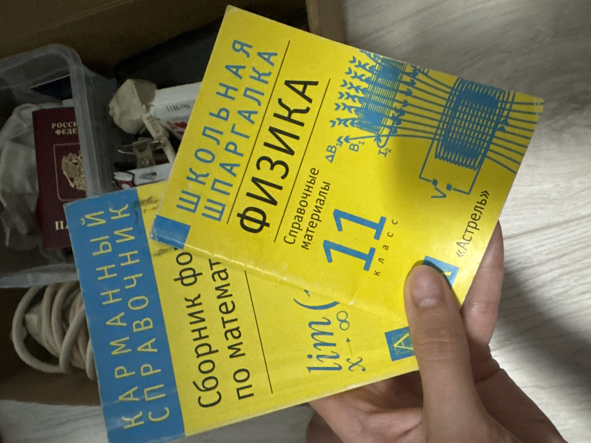 The image shows two Russian school reference books being held in someone's hand. One is a yellow-covered physics reference book for 11th grade, published by Astrel. The title on the cover is "School Cheat Sheet: Physics." The other is a blue-covered reference book on mathematics, titled "Pocket Reference Book: Collection of Formulas in Mathematics." They are being held over a surface with a wood grain pattern. In the background, there is a box with items including a Russian passport. There is no specific indication of Izhevsk, Russia.