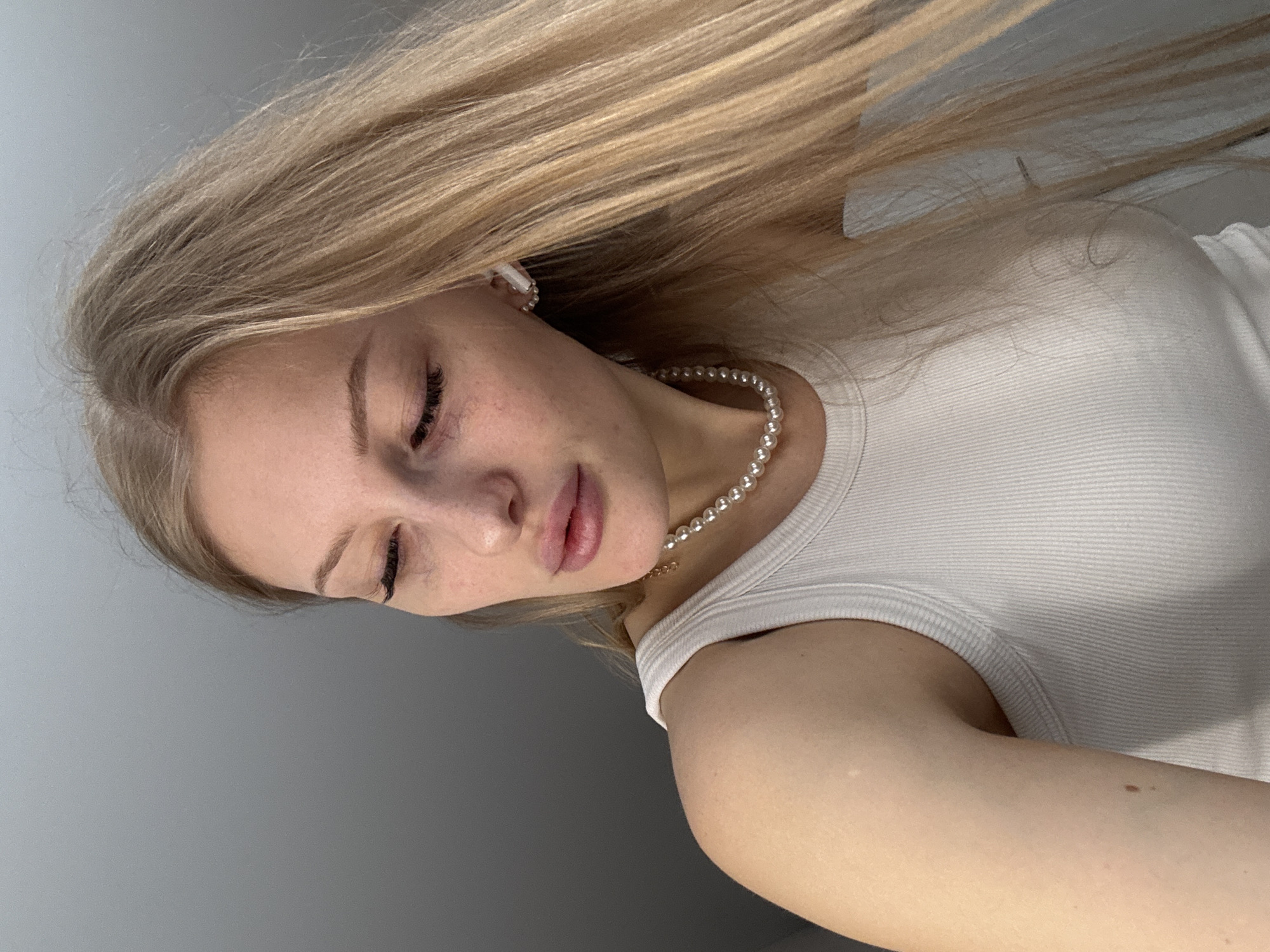 A young woman with long blonde hair is pictured in an indoor setting. She is wearing a white top and a pearl necklace, and has a white earbud in her ear. The background appears to be a plain gray wall. It's not possible to identify the location as Perm, Russia, or any other specific city without further information.