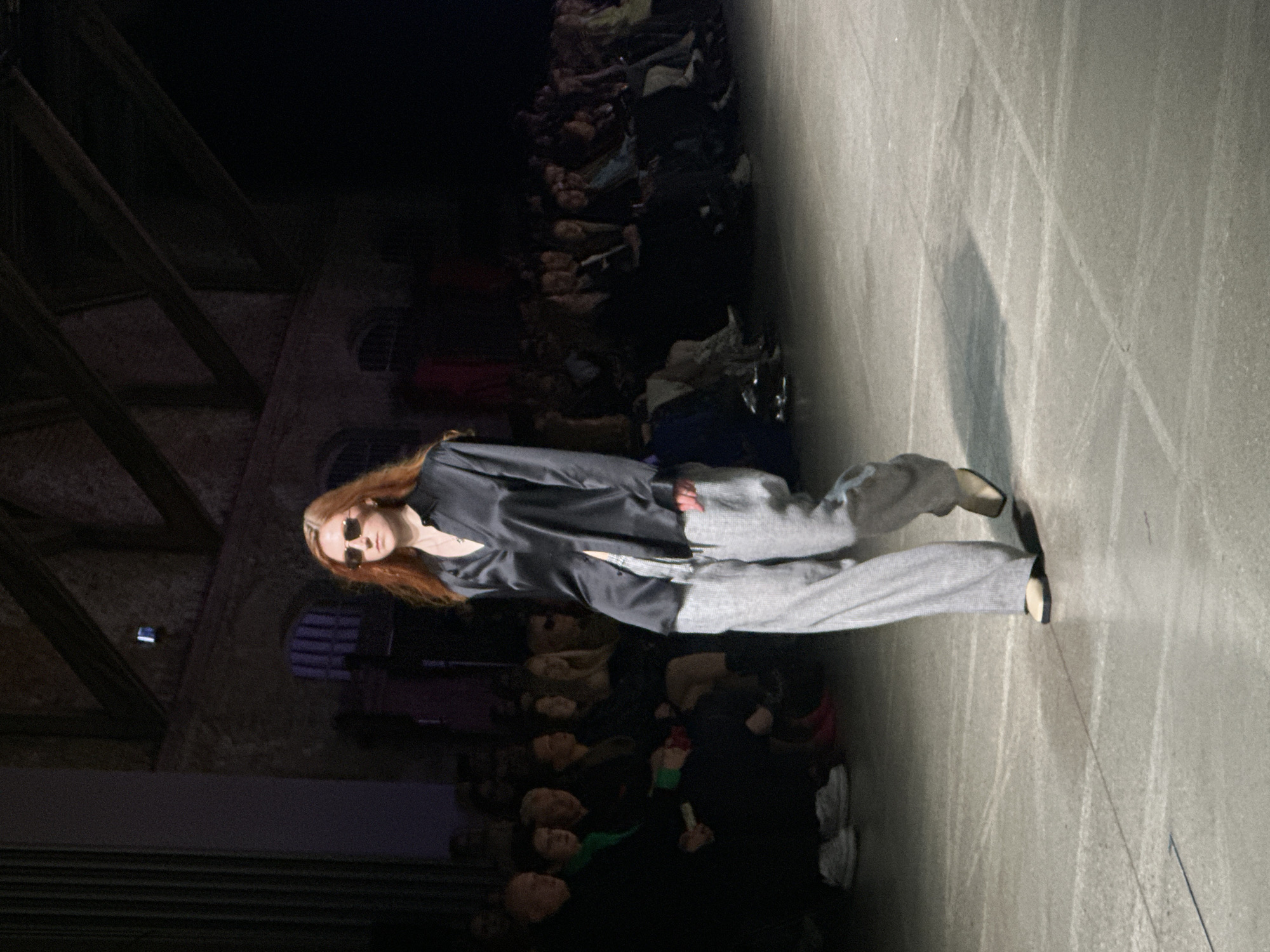 A fashion model with red hair and sunglasses is walking on a gray runway during a fashion show. She is wearing a dark top and light gray pants. Audience members are seated on either side of the runway. The show appears to be taking place indoors in an older building with exposed rafters and arched windows. The location is Riga, Latvia.