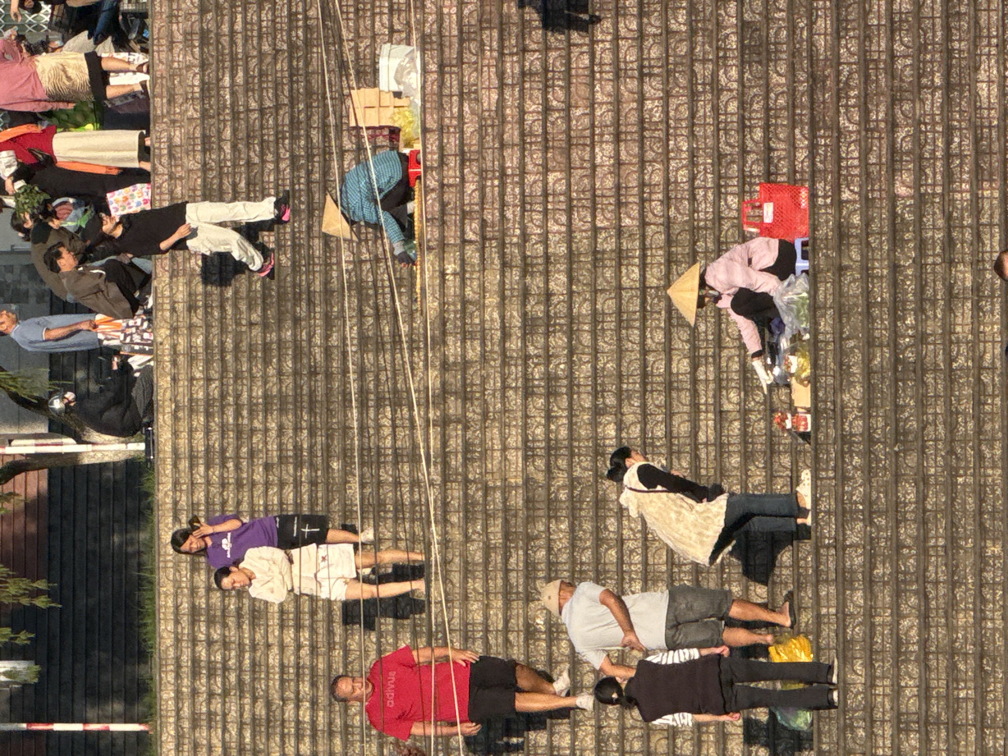 The image captures a wide shot of a terraced outdoor stairway in Da Lat, Vietnam, with several individuals engaged in various activities. The stairway is constructed from stone and has multiple levels.
In the upper portion of the image, on the top tier, a group of people are visible. One person in a blue shirt walks down the stairs, while others stand and interact.
Further down the stairs, a woman in a purple shirt and a woman in a light-colored jacket stand together. Below them, a man in a red shirt and black shorts walks down the stairs, accompanied by a person in a grey shirt and denim shorts.
On a lower level, two individuals are visible. One, wearing a vest and striped shirt, stands to the left. To the right, a person in a beige jacket and dark pants is bending over.
On a mid-level of the stairs, a person wearing a conical hat and blue and white striped clothing sits with their back to the camera, appearing to be selling items. A red bag is placed next to them. Further down and to the right, another person wearing a conical hat and a pink top is seated, also appearing to be selling goods. A red bag is situated to their right.
The environment suggests a public space, possibly a market area or a tourist attraction, given the presence of vendors and the style of clothing. The lighting indicates it is daytime, with shadows cast on the stone steps.
