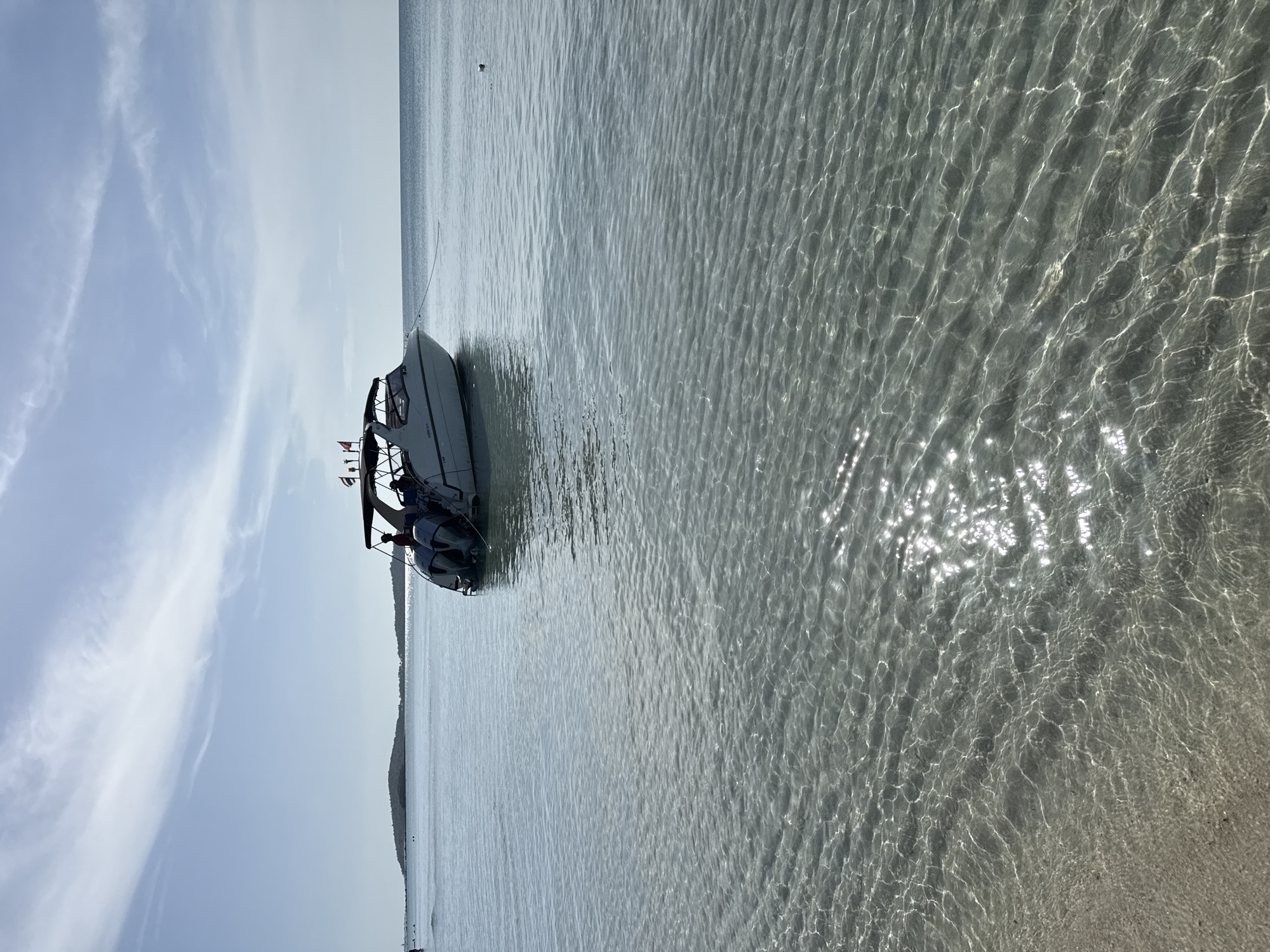 The image shows a white speed boat with two outboard motors anchored in crystal clear shallow water off the coast of Ko Samui District, Thailand. The boat has a canopy and several people are visible on board. The sun is shining, creating shimmering reflections on the water's surface. In the background, a distant island is visible, as is a calm sea and a partly cloudy sky. The sandy seabed is visible through the translucent water.