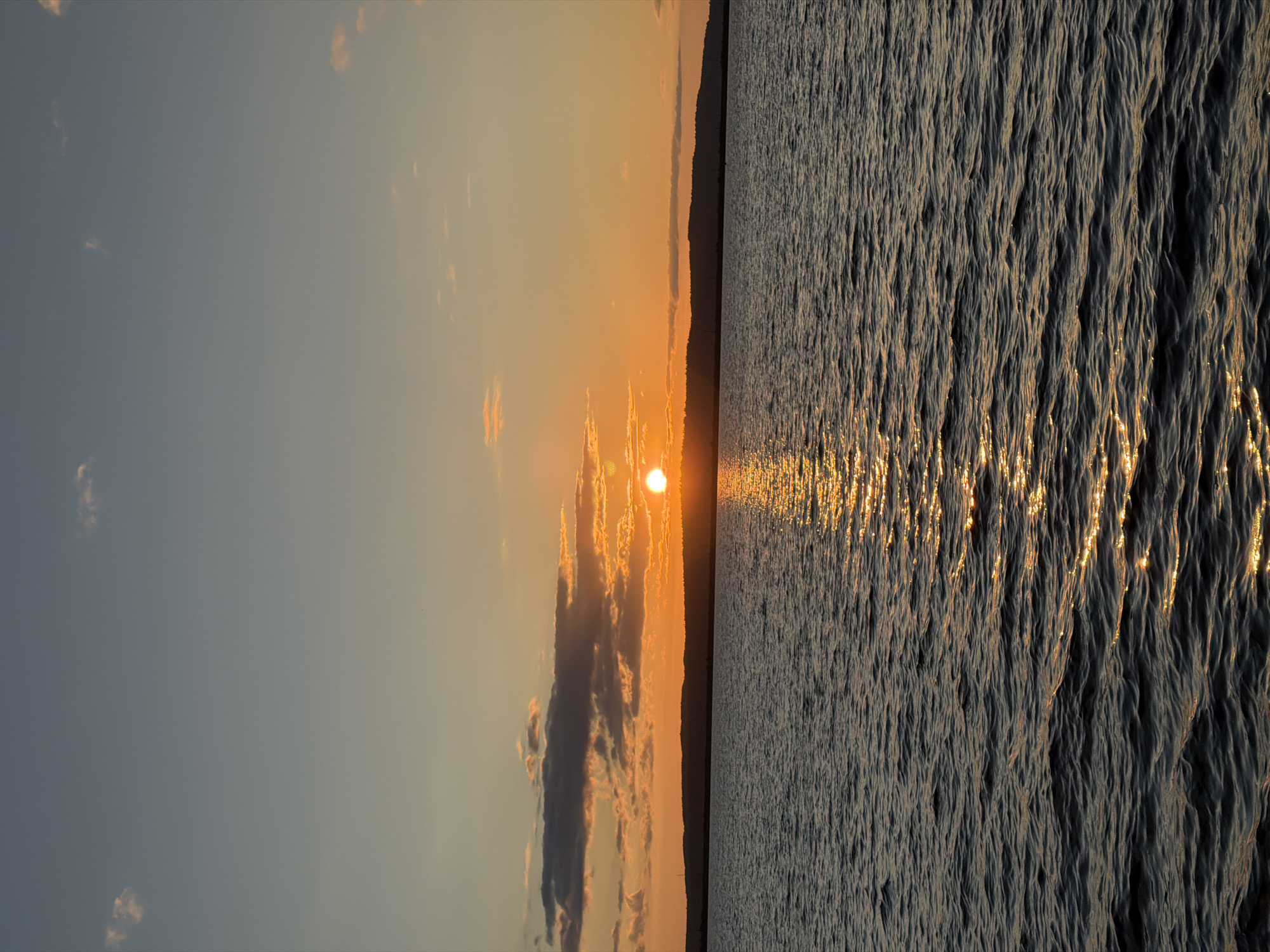 A serene sunset over a body of water, likely a reservoir given its expanse. The sun, a bright orb, is positioned just above the horizon line, casting a shimmering golden path across the rippling surface of the water. The water itself appears dark, with gentle waves creating texture and reflecting the warm hues of the sunset. In the background, a silhouetted treeline forms the horizon, suggesting a natural landscape. The sky transitions from a pale blue at the top to warmer oranges and yellows near the horizon, with a few scattered clouds adding depth and visual interest. The overall impression is one of tranquility and natural beauty. The location is identified as Chaikovsky, Russia.
