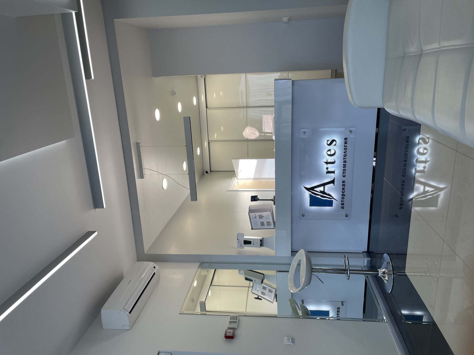 The image depicts the interior of a dental clinic, presumably located in Izhevsk, Russia, as indicated by the Cyrillic script on the signage. The clinic's reception area is prominent, featuring a high, white reception desk with the clinic's logo "Artes" prominently displayed. A chrome bar stool is positioned in front of the desk.
The surrounding environment is clean and modern, with white walls and glossy tile flooring. Linear LED lighting fixtures are integrated into the ceiling, along with round recessed lights. A white air conditioning unit is mounted on the wall. A mirror partially reflects the reception desk and the surrounding area.
The overall impression is a bright, professional, and modern medical environment.
