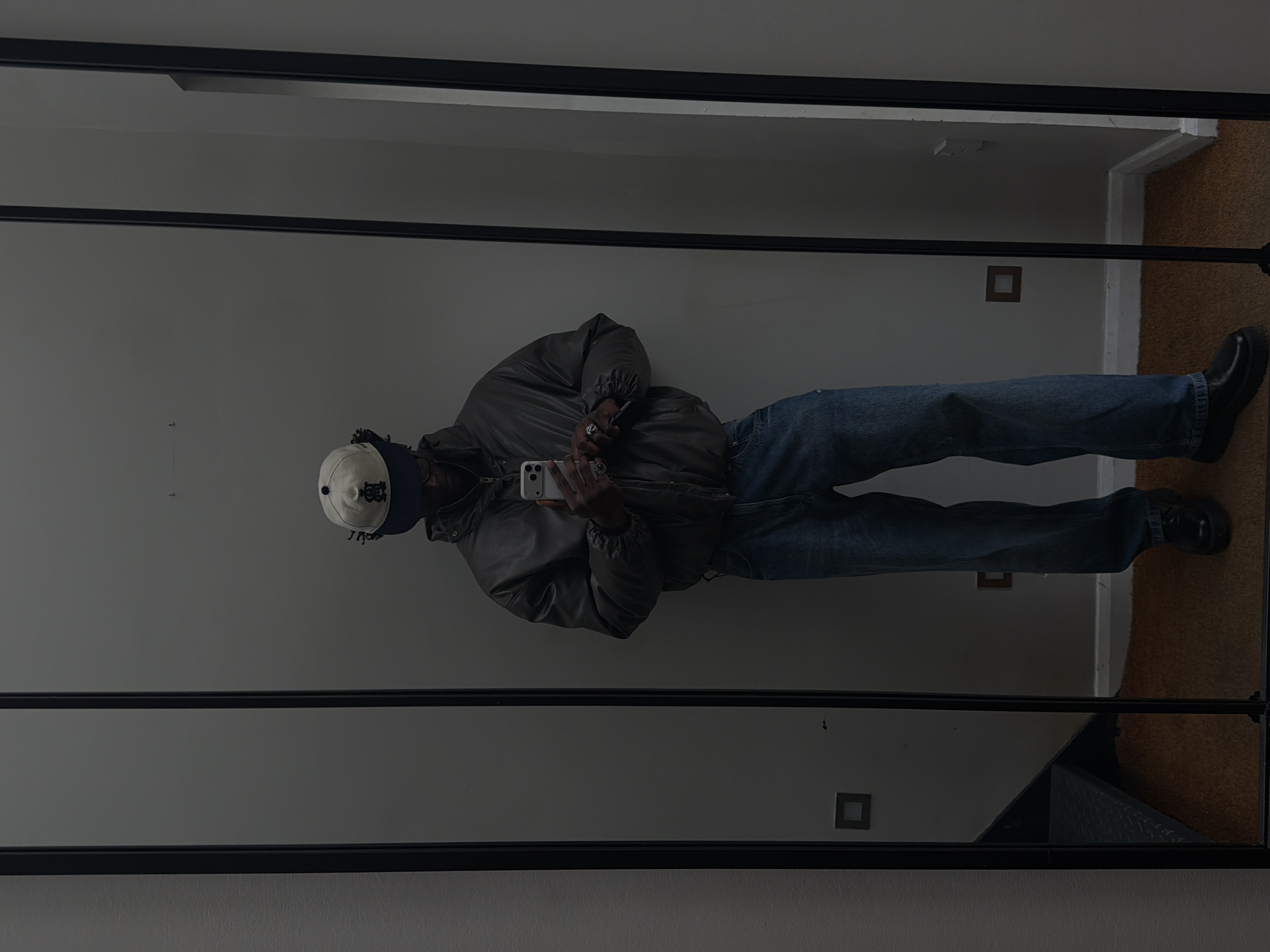 A person is reflected in a large mirror with a black frame. The person is wearing a light-colored baseball cap, a dark blue beanie, a brown puffer jacket, blue jeans, and black shoes. They are holding a smartphone in their hands, presumably taking a selfie. The background behind the reflection includes a white wall with electrical outlets and a section of carpeted floor. The image is indoor, and the location cannot be determined based on the provided information.