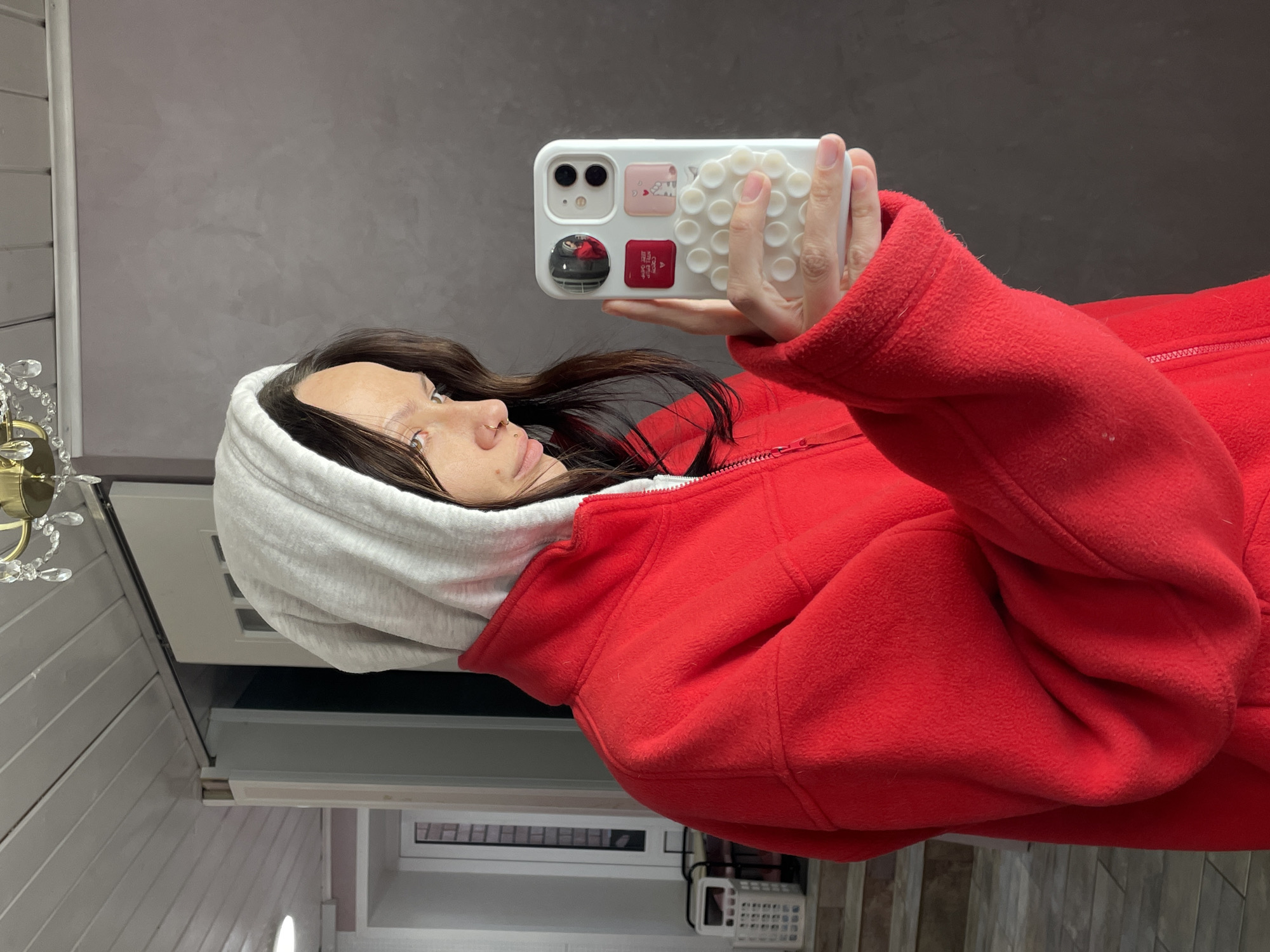 A woman in a red hooded fleece jacket takes a selfie with a white cellphone. The cellphone has a white case with raised suction cups and several stickers. The woman has dark brown hair and is partially obscured by the hood. The background includes a light gray wall, a white wall with vertical panels, and a chandelier. It is not possible to definitively determine the location of the scene as Izhevsk, Russia.