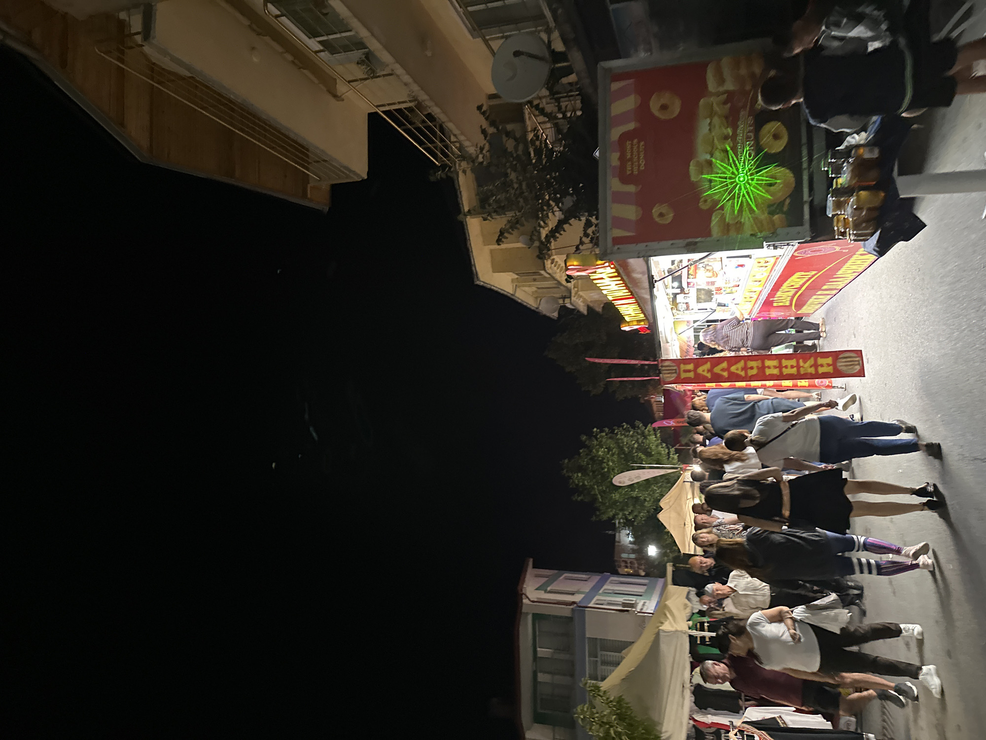 The image depicts a night scene at what appears to be a street market or festival in Nedelino, Bulgaria. A crowd of people is walking along a street lined with market stalls and food vendors.
In the foreground, a food truck with a prominent red and yellow striped awning is visible. The truck's side displays an advertisement for "THE MOST DELICIOUS DONUTS" with images of donuts and a green laser-like starburst graphic. In front of the truck, a table displays several jars of what appear to be preserves or honey. Two young boys are standing near this table.
To the left of the food truck, there are market tents with clothing items displayed. Several people are browsing these stalls. The street is paved, and the overall lighting is dim, suggesting it is nighttime. Buildings are visible on the sides of the street, with balconies and windows. The sky above is dark, with some faint atmospheric lights or reflections.
Further down the street, more people are walking and interacting. The general atmosphere suggests a lively evening event. The signs and lettering on the stalls are likely in Bulgarian, reinforcing the location.