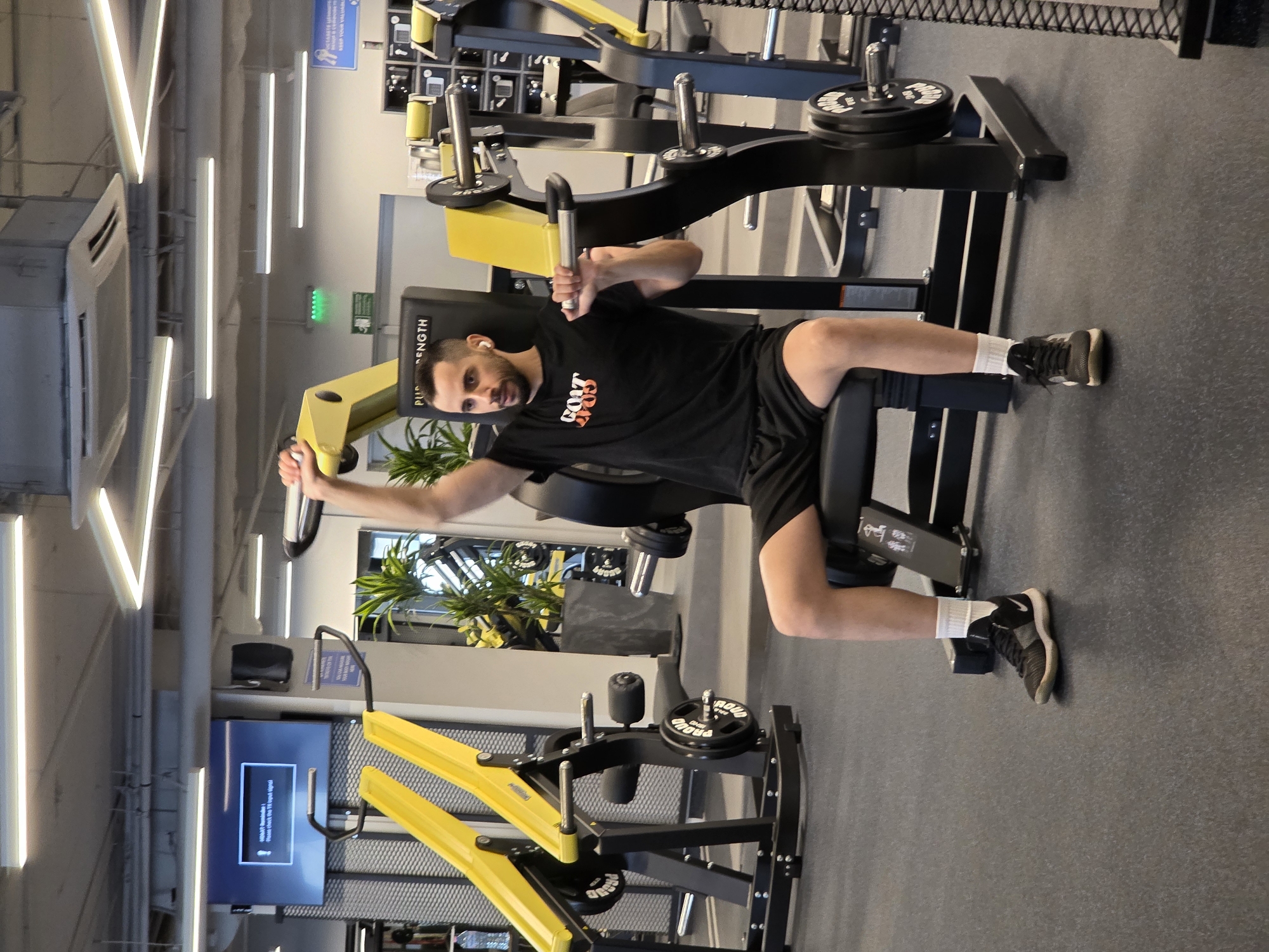 The image captures a man posing in a modern gym environment, identified as being in Plovdiv, Bulgaria. The man, likely in his 20s or 30s, has a beard and short dark hair. He is wearing a black t-shirt with "GOAT GYM" printed on it, black shorts, white socks, and dark sneakers. He also has wireless earbuds in his ears. He is seated on a yellow and black gym machine, resembling a chest press or pec deck, with his arms extended and hands gripping the handles, looking directly at the camera with a neutral expression.
The gym features bright artificial lighting from linear LED fixtures on the ceiling and a dark grey rubberized floor. Various other black and yellow gym machines are visible in the background, some with weight plates marked "PROUD." A large ceiling-mounted air conditioning unit and green potted plants are also part of the decor.
On a wall to the left, a flat-screen television displays an error message: "Display not supported! Please check the TV input signal." A blurry blue sign with text, likely in Bulgarian, is also present in the background. The text "STRENGTH" is visible on the padded part of the machine the man is using. The overall setting is that of a contemporary and well-equipped fitness center.