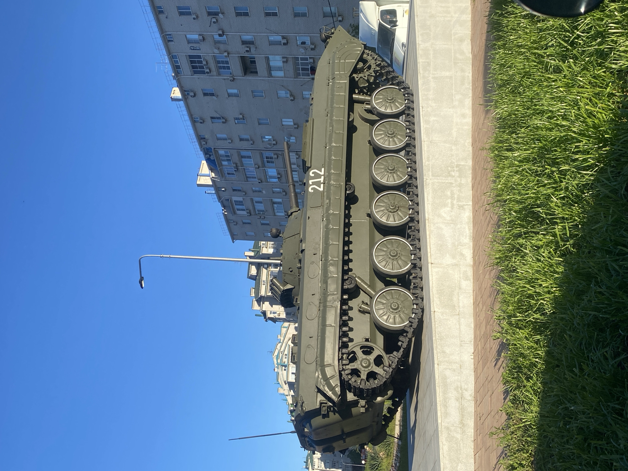 The image displays a green military tracked vehicle, possibly an Infantry Fighting Vehicle (IFV) or Armored Personnel Carrier (APC), positioned outdoors on a light-colored paved surface. The vehicle has the number "212" painted in white on its side. To its right, there is a brown brick path bordering a strip of vibrant green grass.
In the background, several multi-story residential buildings with numerous windows and visible air conditioning units rise under a clear blue sky, suggesting an urban or suburban environment. The buildings appear to be made of light-colored brick or concrete.
The scene is brightly illuminated by natural sunlight, indicating it is daytime, likely mid-day or afternoon, as evidenced by the distinct shadows cast by the vehicle and a tall, slender streetlamp on the left. A portion of a white truck is visible behind the military vehicle, nestled among the background buildings.
No people are visible in the image, and no specific activity or event is taking place beyond the static display of the military vehicle, which appears to be a monument or exhibit.